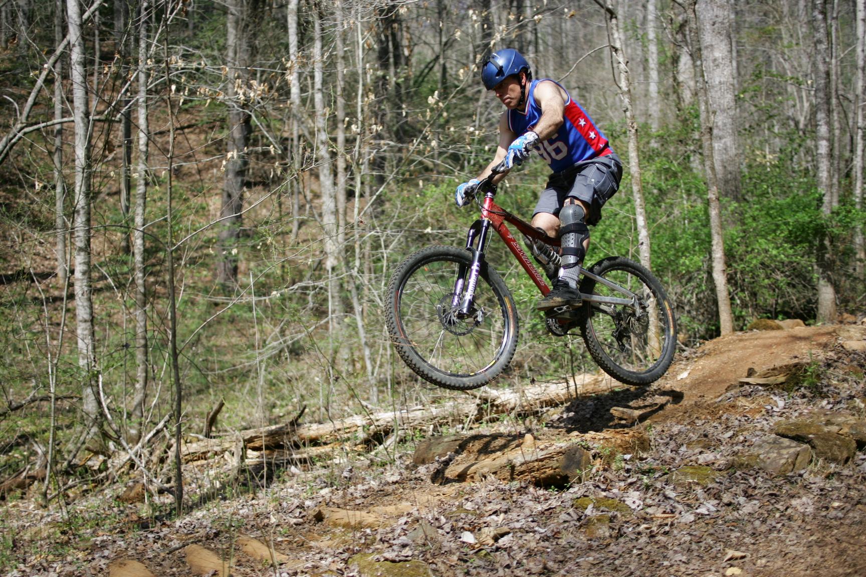 A mountain biker in protective gear jumps off a rocky trail in a forested area, showcasing a dynamic action shot with trees and greenery in the background. Dark Mountain Trail mountain bike trail.