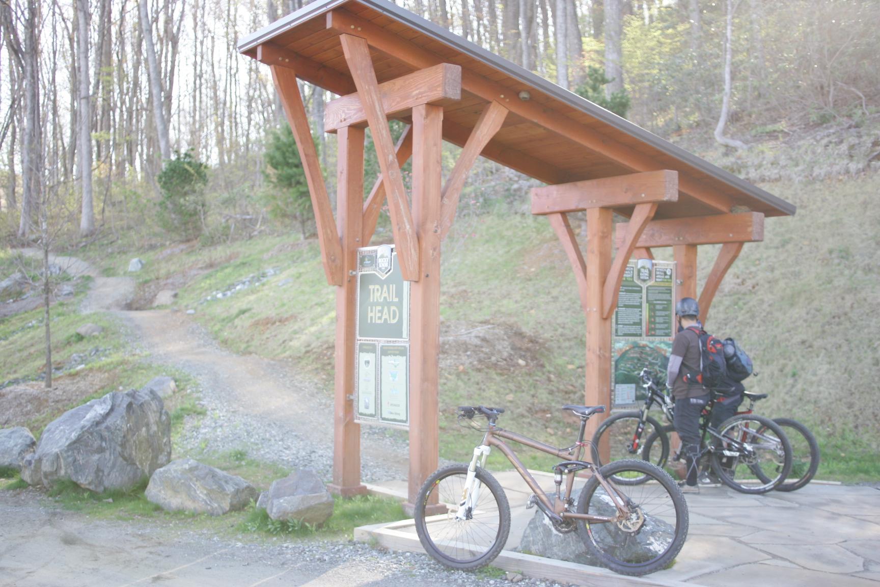 A mountain biker stands next to a trailhead sign at the start of a biking trail, with two bicycles parked nearby. The surroundings feature a grassy hill and rocky terrain, surrounded by trees in a forested area. The sign provides information about the trail, including maps and guidelines for bikers. Rocky Knob Park mountain bike trail.