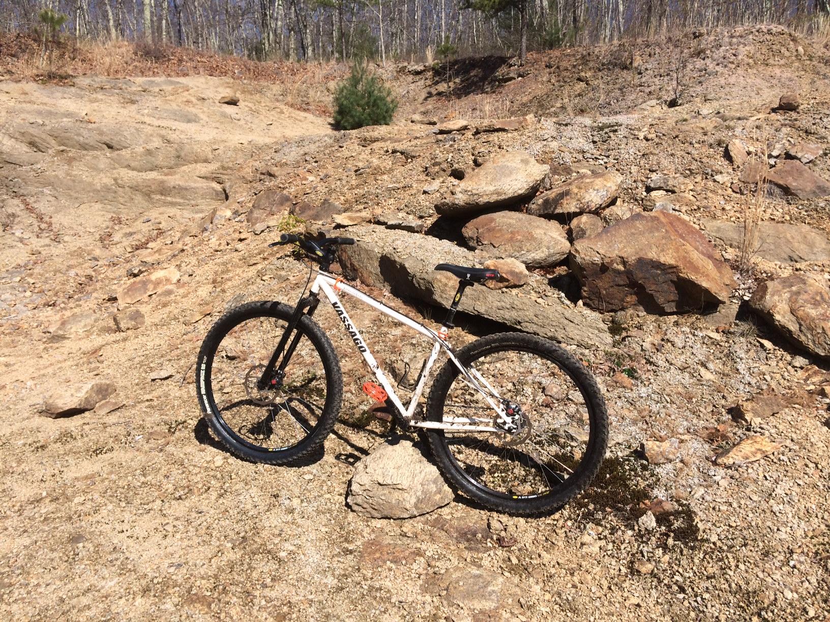Vassago Jabberwocky: A mountain bike rests on rocky terrain under clear blue skies, surrounded by sparse vegetation and exposed earth. The bike has a white frame and black tires, positioned next to a large rock, indicating a trail or off-road setting.