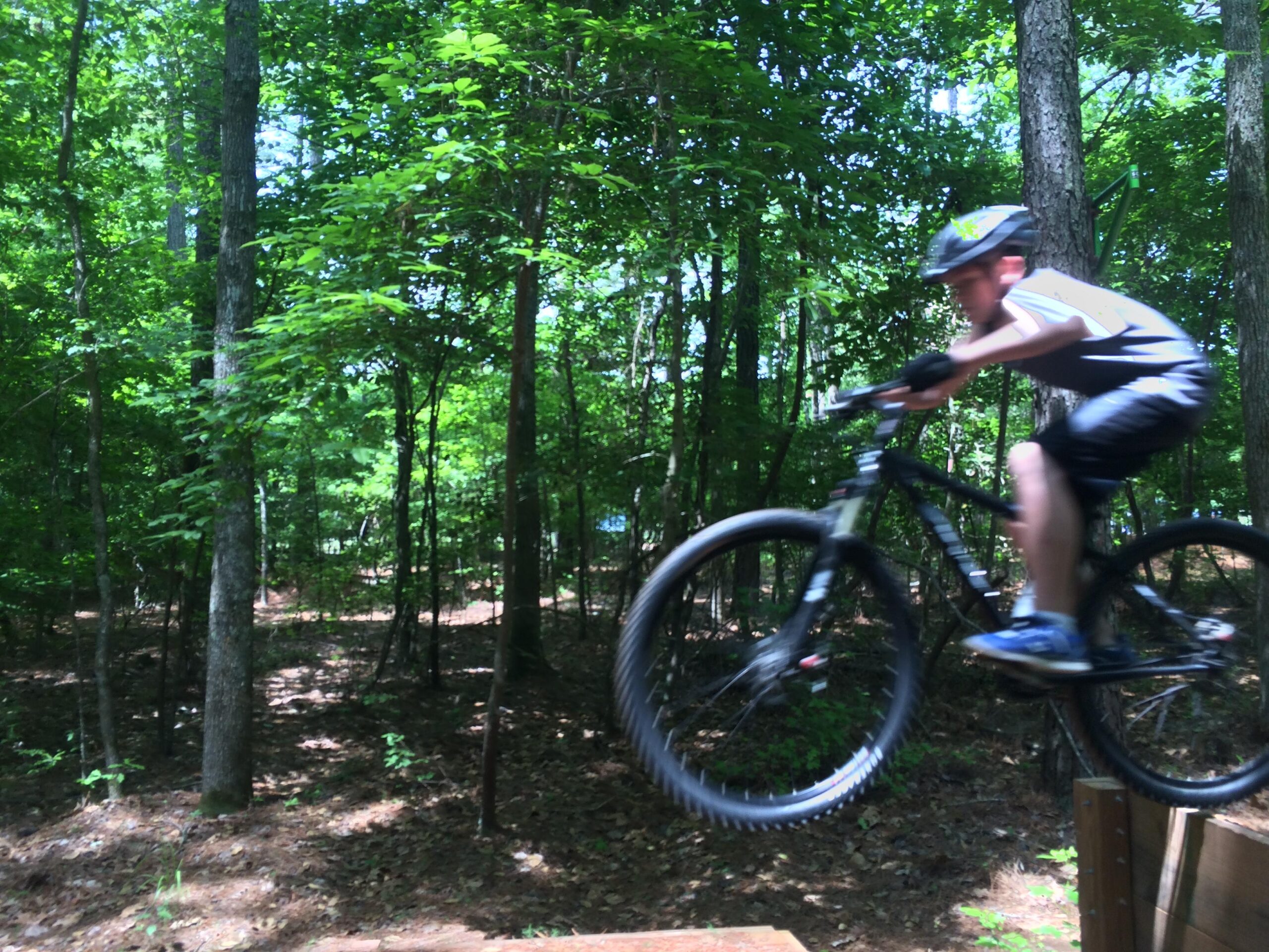 Gary Fisher Rig 29er: A child riding a mountain bike jumps off a ramp, surrounded by lush green trees in a wooded area. The rider is captured in mid-air, showcasing dynamic movement and excitement.