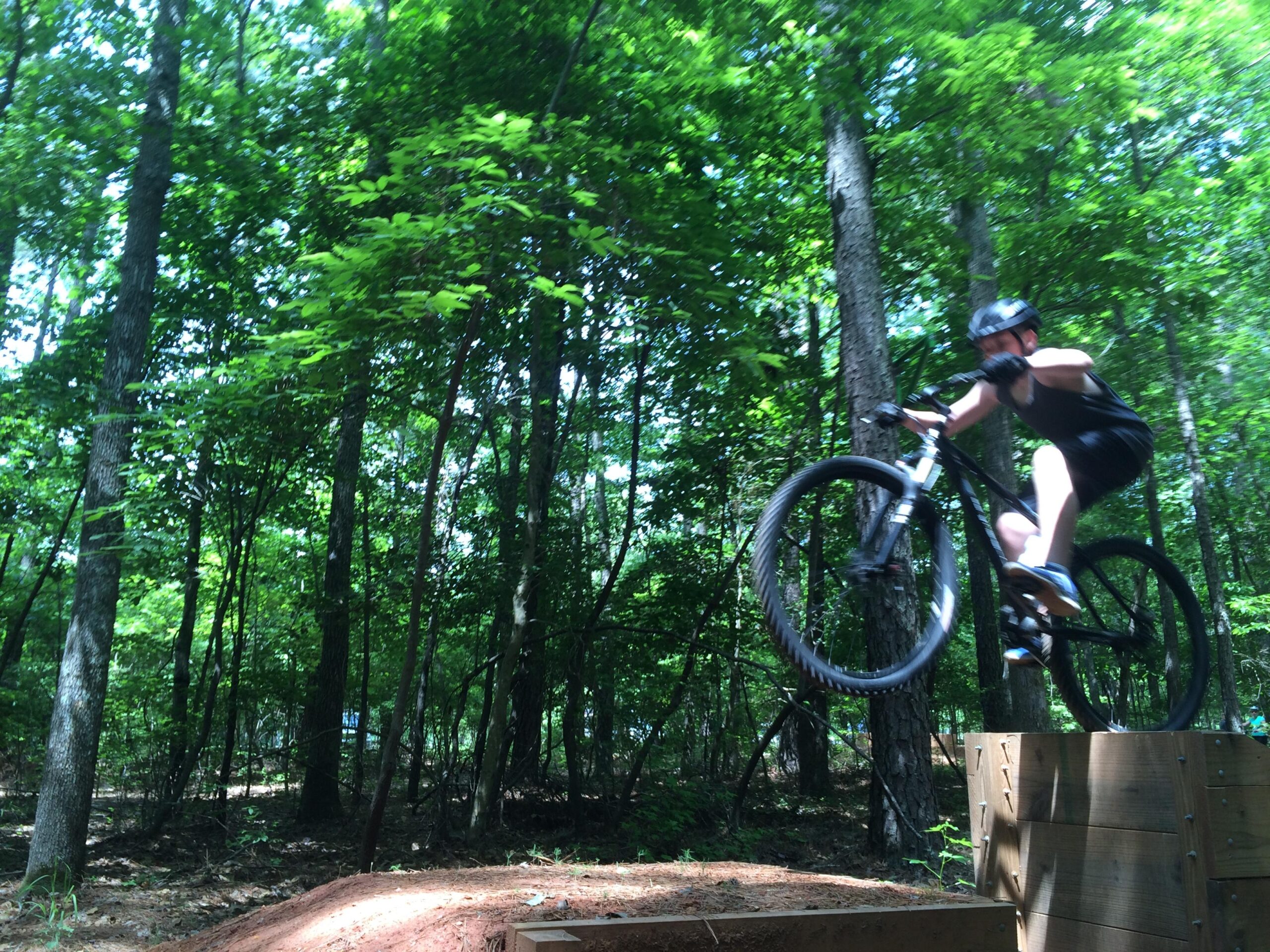 Gary Fisher Rig 29er: A young person performing a jump on a mountain bike off a wooden ramp in a densely wooded area, surrounded by lush green trees. The rider is wearing a helmet and appears focused as they catch air above the ramp.