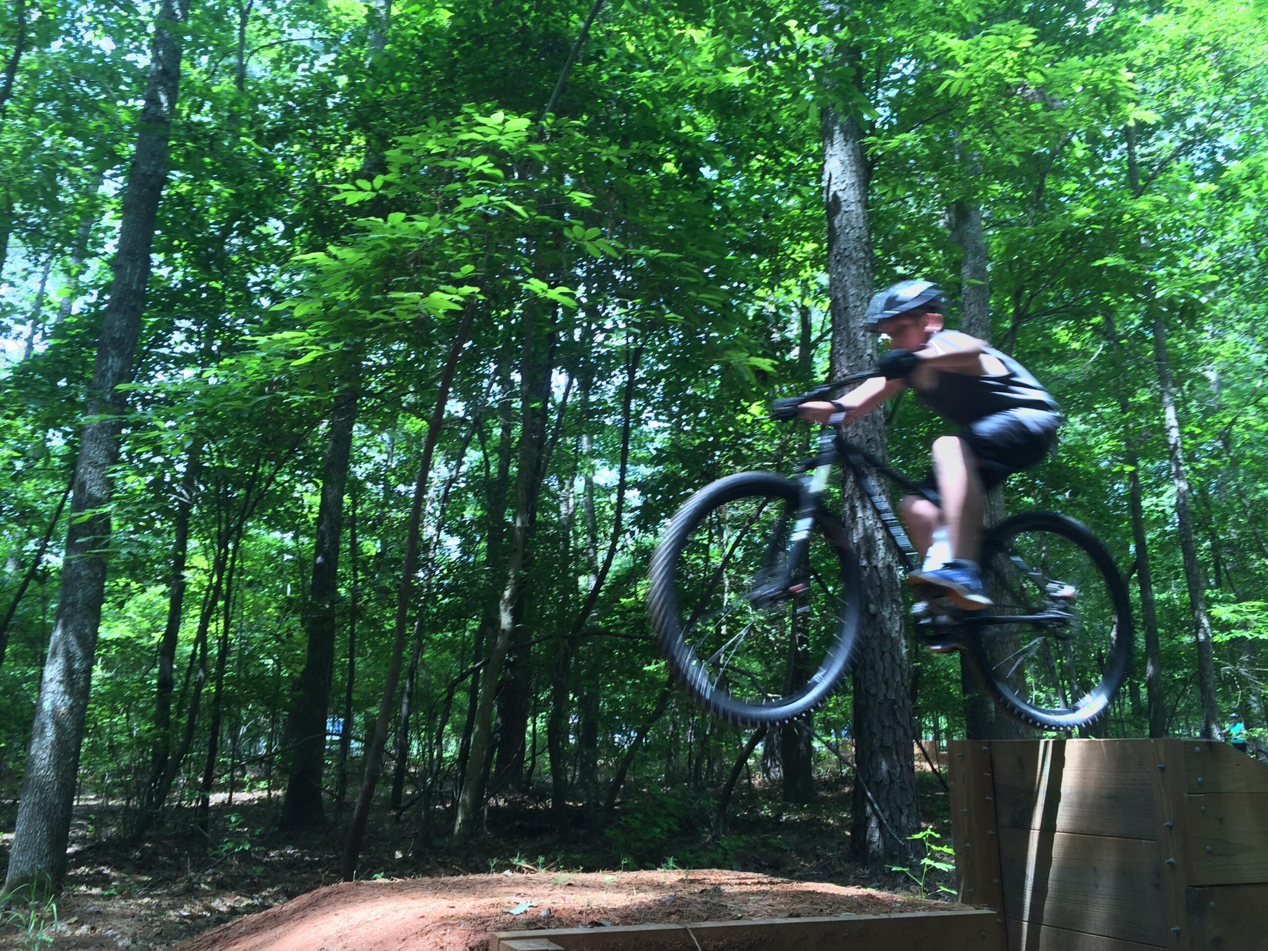 Gary Fisher Rig 29er: A young boy in a helmet jumps off a wooden ramp while riding a mountain bike through a wooded area, surrounded by lush green trees.