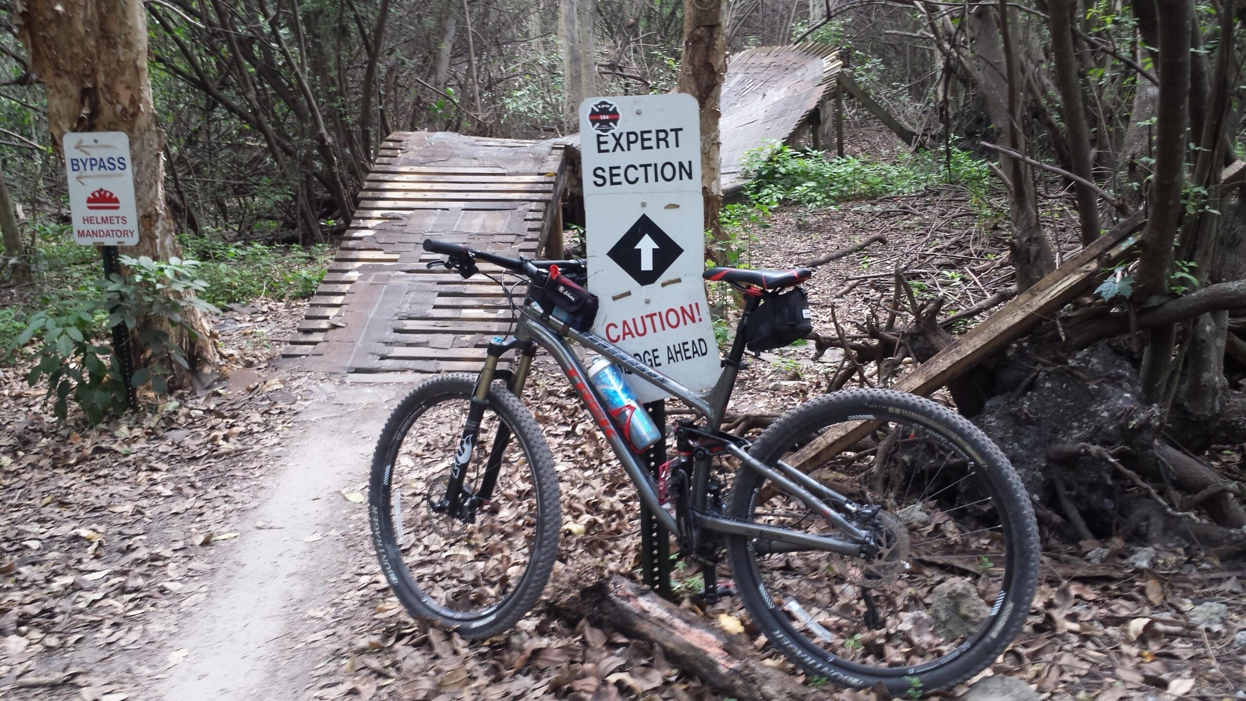 Trek Fuel EX 8 29: A mountain bike parked beside a trail sign that reads "Expert Section" with a caution symbol and an arrow pointing forward. There's another sign indicating "Bypass" and that "Helmets Mandatory," along with a wooden ramp visible in the background surrounded by dense foliage and fallen leaves.