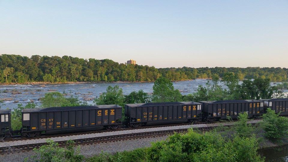 A train of black freight cars labeled with "CSX" runs along a railway beside a river, with lush green trees and a rocky riverbank in the background. The scene is set in the early morning or late afternoon light, creating a peaceful landscape. Buttermilk mountain bike trail.