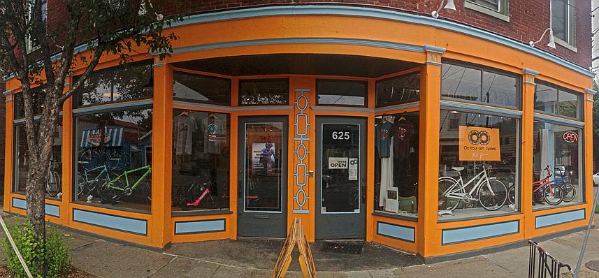A vibrant bike shop exterior featuring large windows showcasing colorful bicycles and merchandise. The building is painted in orange and blue hues, with signage indicating the shop