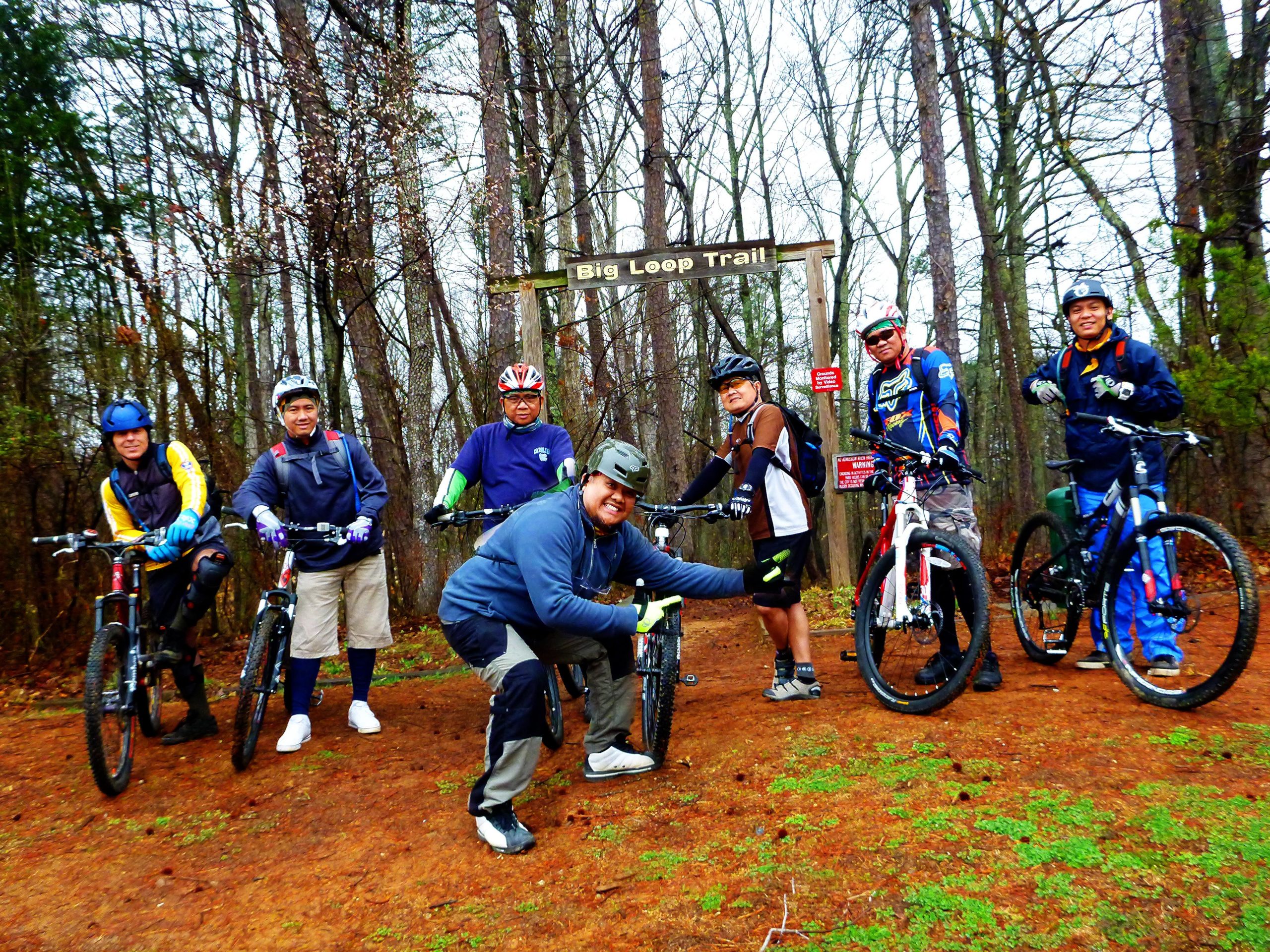 A group of six mountain bikers poses at the entry of the Big Loop Trail, surrounded by tall trees. The riders, wearing helmets and colorful biking gear, stand with their bicycles on a dirt path. The atmosphere is lively, showcasing camaraderie among friends ready for an outdoor adventure. Big Loop mountain bike trail.