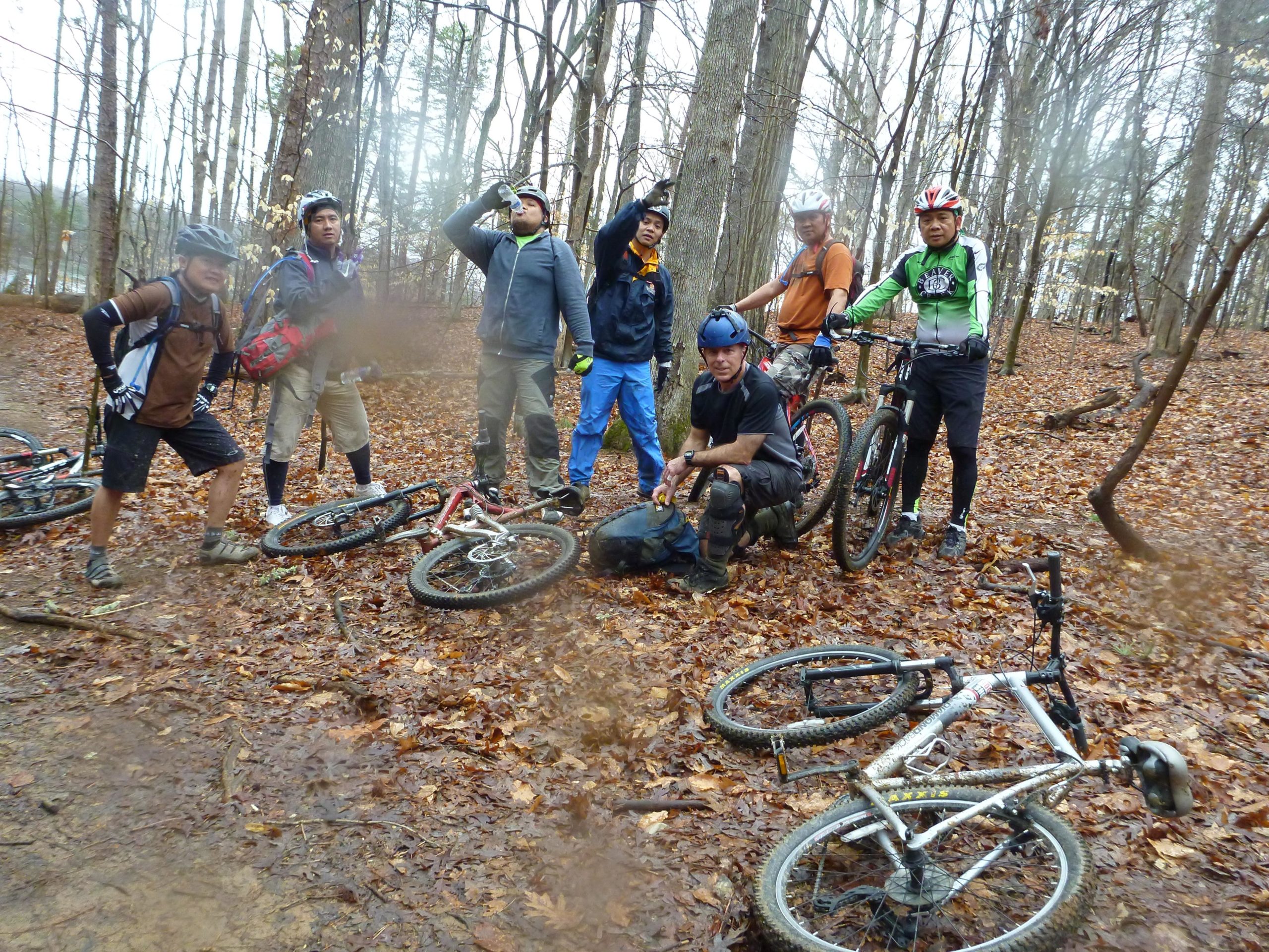 A group of seven mountain bikers gathered in a wooded area covered with fallen leaves. Some are standing near their bikes, while one person kneels beside a backpack. The bikers wear helmets and various cycling gear, and are friendly and engaged with each other in a natural outdoor setting. The atmosphere appears casual and relaxed, with trees and nature surrounding them. Big Loop mountain bike trail.