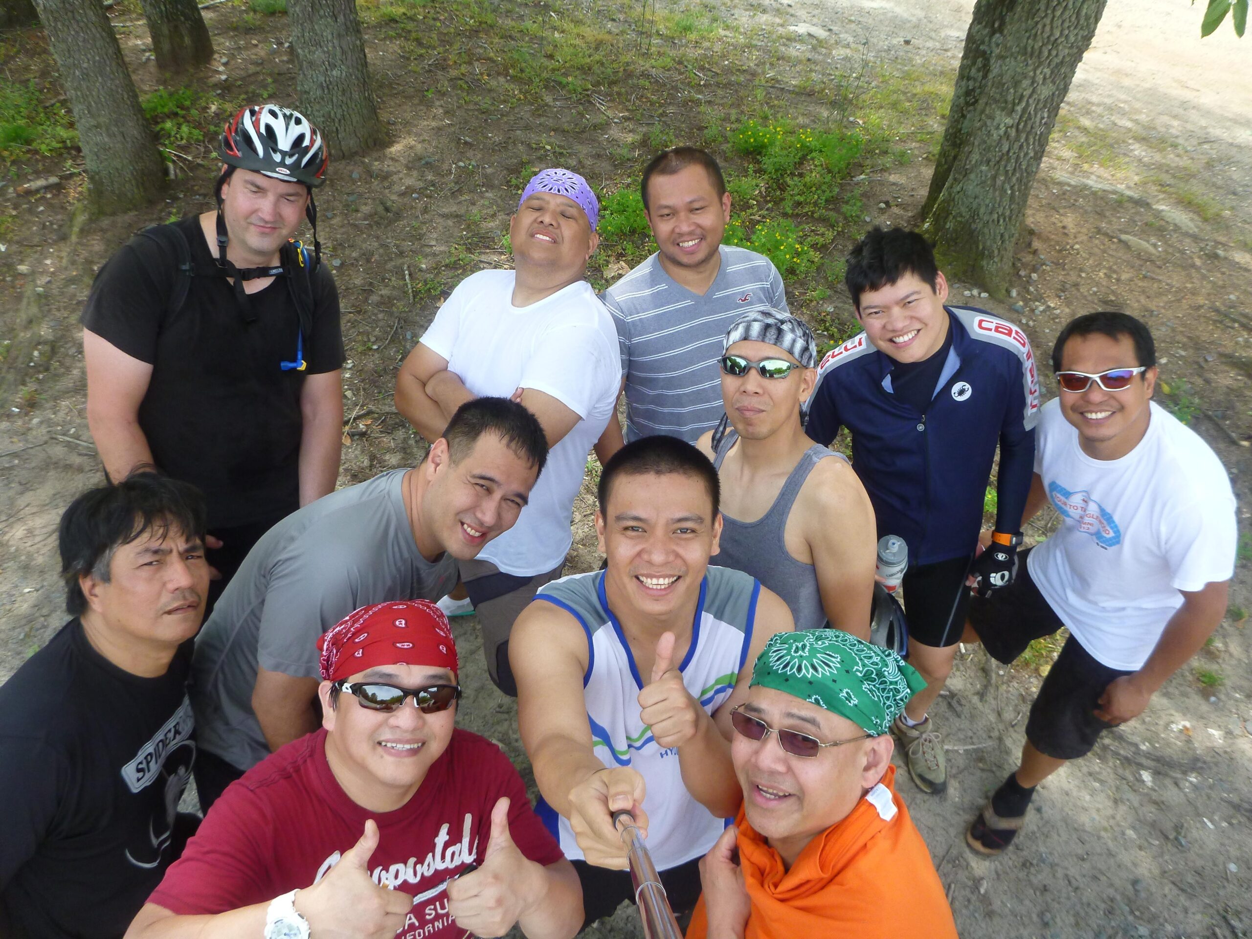 A group of eleven men posing for a group selfie outdoors. They are wearing casual sports attire, including helmets and bandanas, and are smiling as they enjoy a day out. The background features trees and a natural setting. Hobby Park mountain bike trail.