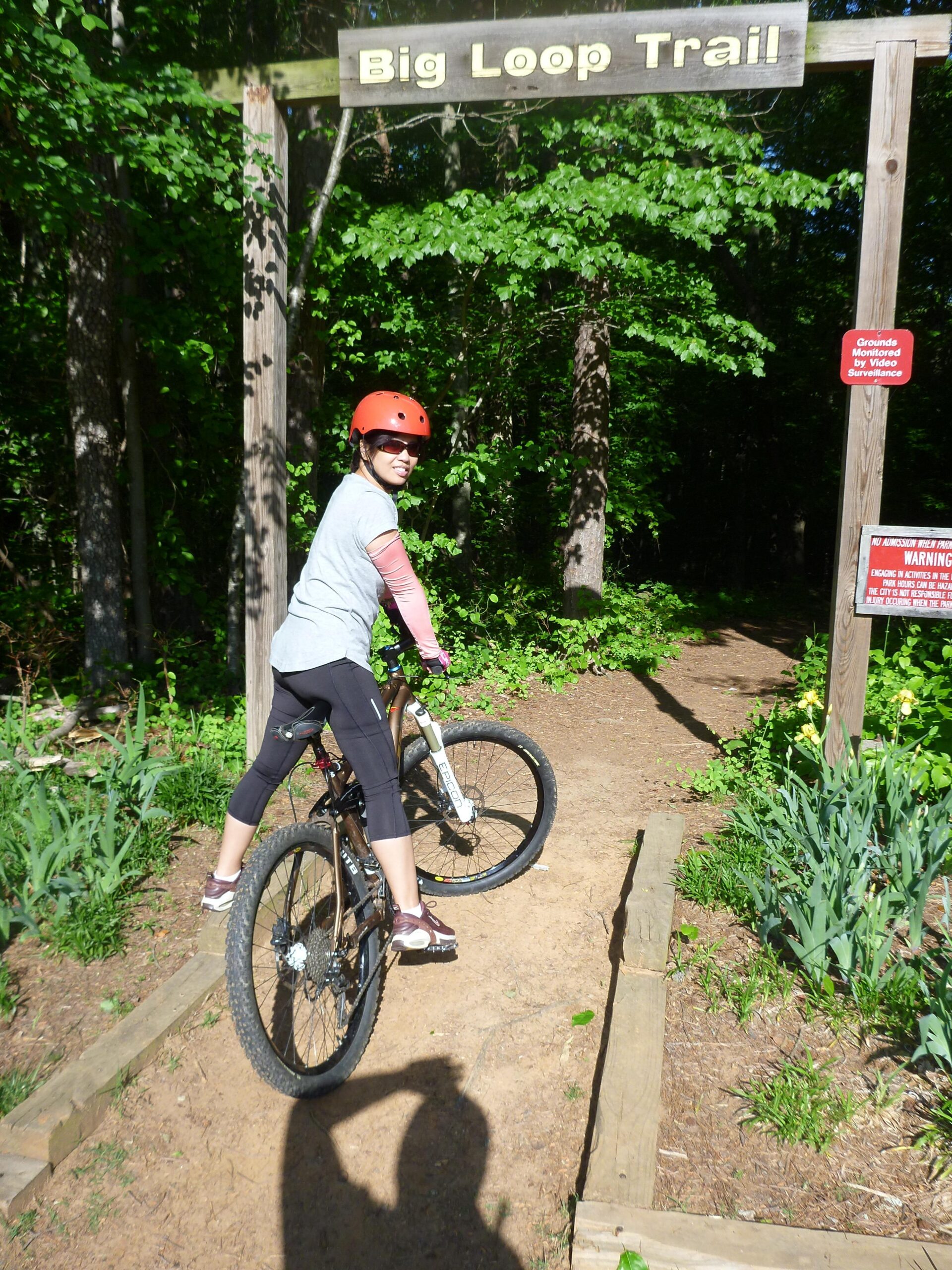 A person on a mountain bike pauses at the entrance to the Big Loop Trail, smiling while wearing a helmet and sporty clothing. The trail entrance is marked by a wooden sign with the name "Big Loop Trail," surrounded by lush green foliage and dirt path leading into the trail. Big Loop mountain bike trail.