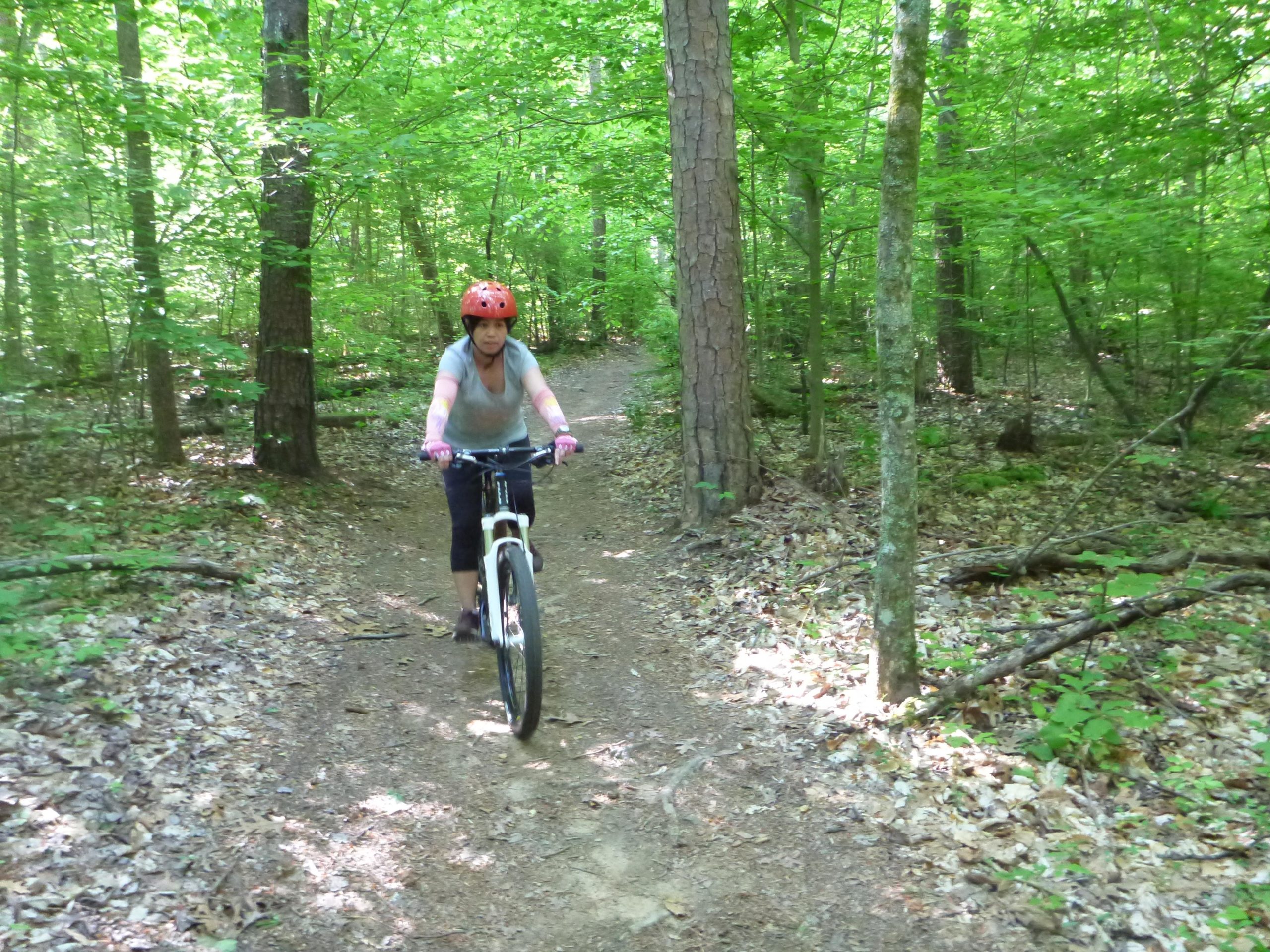 A person riding a mountain bike along a dirt trail surrounded by lush greenery and tall trees in a sunny forest. The individual is wearing a bright orange helmet and has a focused expression while navigating the path. Big Loop mountain bike trail.