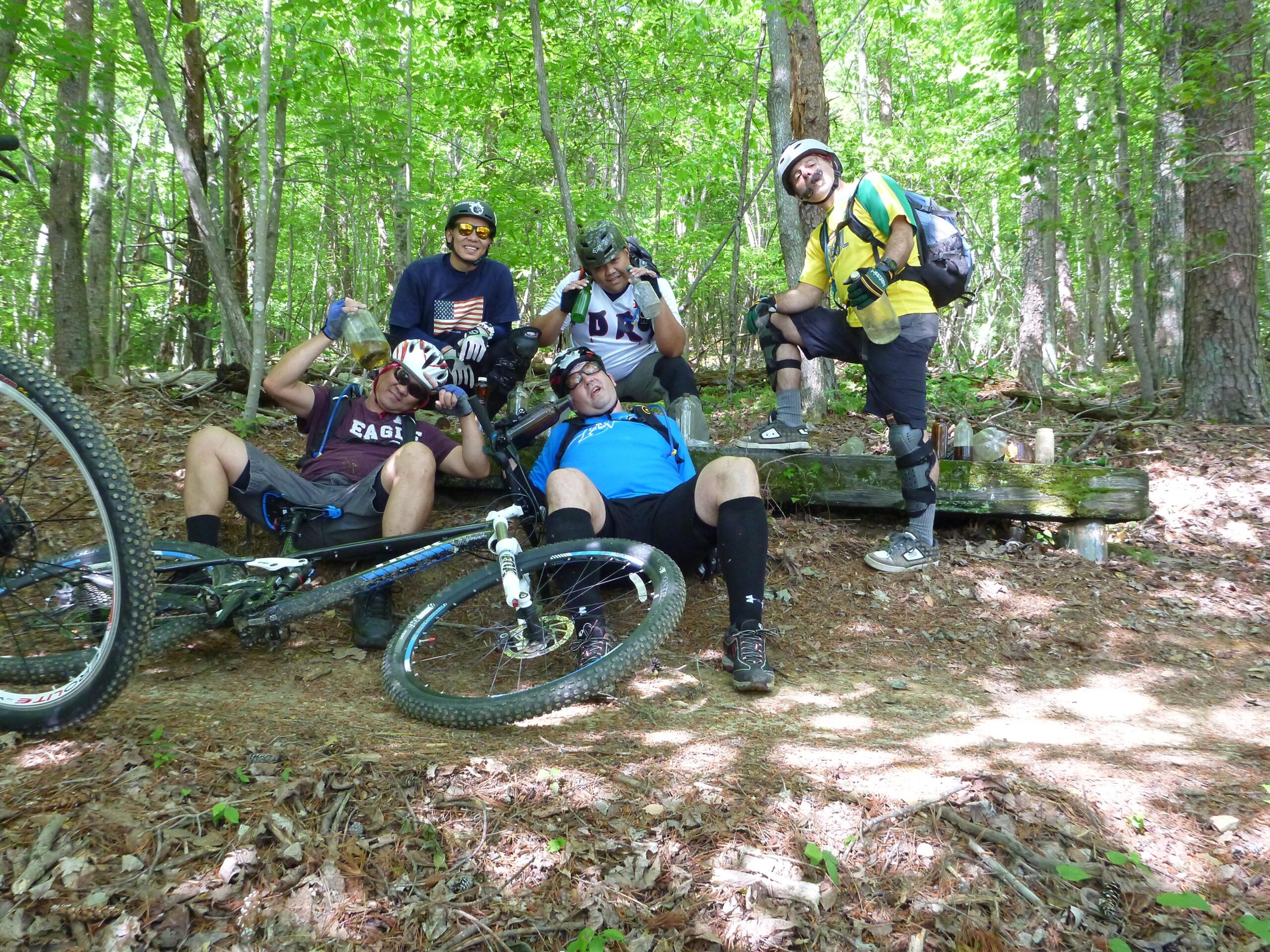 A group of five mountain bikers are posing together in a forested area. They are enjoying a break amidst the green trees, with some sitting on the ground and others standing. One person is lying down next to a bike and holding a bottle, while another strikes a playful pose. All are wearing helmets and casual biking gear. The scene is lively and captures the camaraderie of outdoor biking adventures. Warrior Creek mountain bike trail.