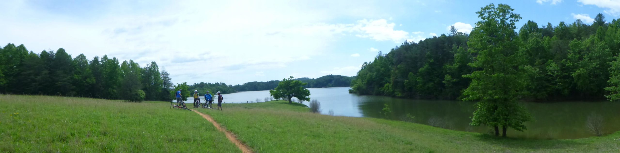 A panoramic view of a serene lake surrounded by lush green trees, with a clear blue sky above. In the foreground, a grassy area leads to a path where a group of cyclists is gathered, enjoying the natural scenery. Warrior Creek mountain bike trail.