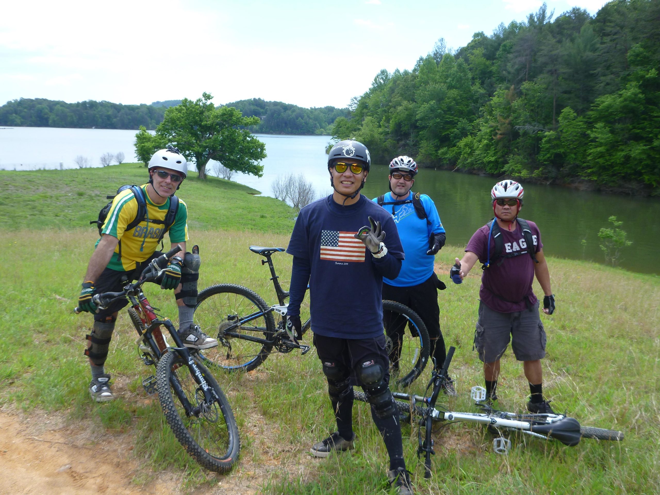 Four mountain bikers pose for a photo on a grassy area by a lake. They are wearing helmets and protective gear. One biker, in the front, flashes a peace sign, while the others smile and give thumbs up. The serene lake is visible in the background, surrounded by lush green trees and hills. Warrior Creek mountain bike trail.