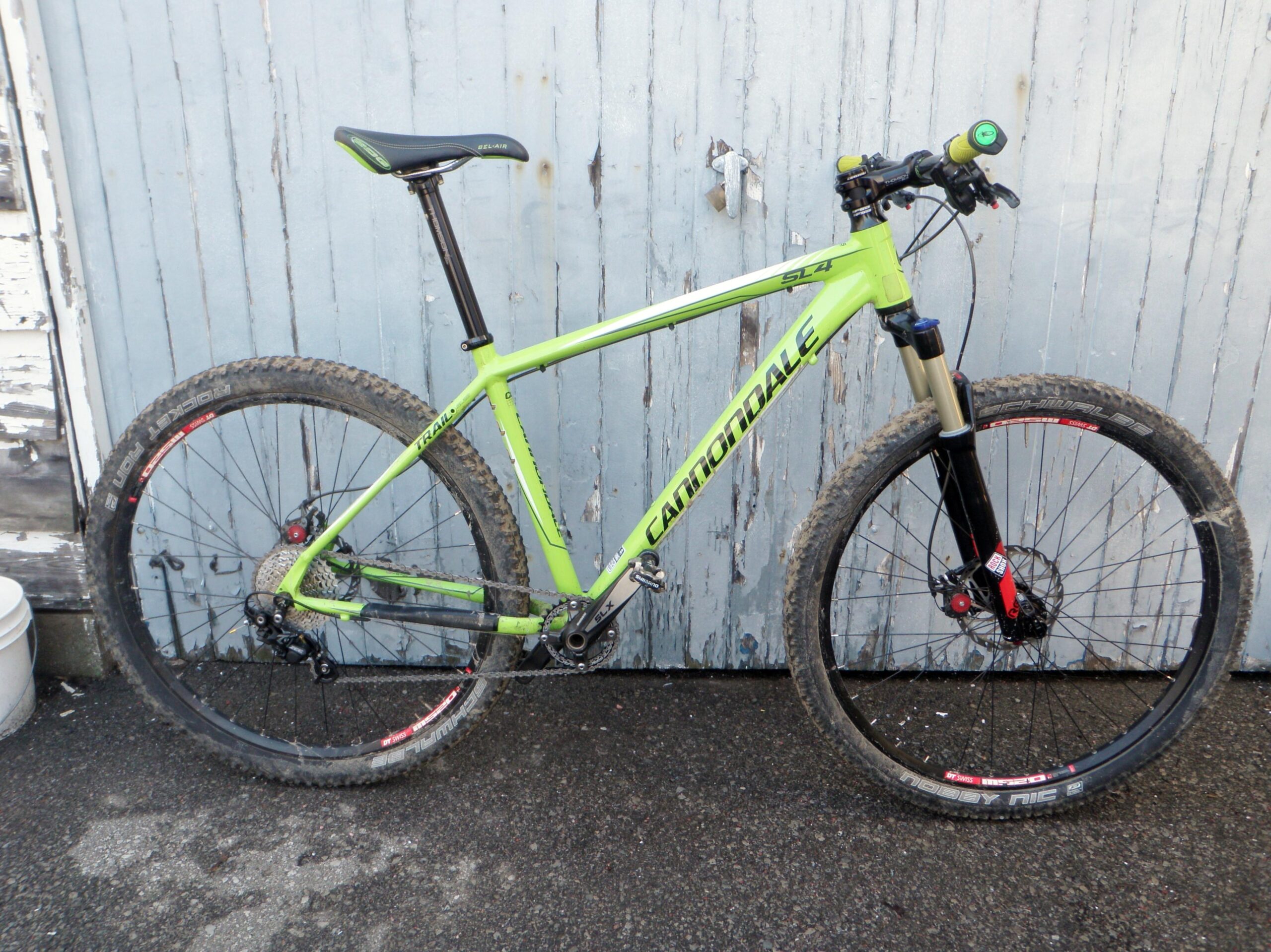 Cannondale Trail SL 29: A bright green Cannondale mountain bike with a black front fork and muddy tires, leaning against a weathered gray wooden wall. The bike features a sleek design, multiple gears, and is equipped for off-road cycling.