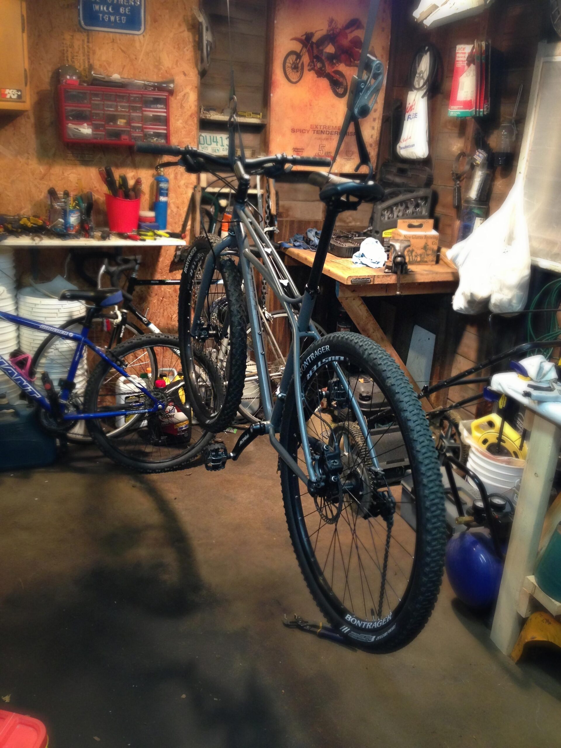 Trek Sawyer: A cluttered garage workspace with two bicycles hanging on a bike rack. Tools and bicycle parts are scattered around, including a workbench with various items and a red toolbox. The walls display signs and posters related to biking and motor sports.