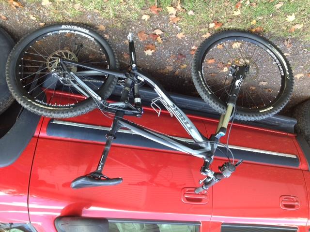 Trek Remedy 7: Mountain bike resting on the roof of a red vehicle, with the wheels facing upwards and fallen leaves scattered in the background. The bike features a black frame, wide tires, and is positioned diagonally across the roof.