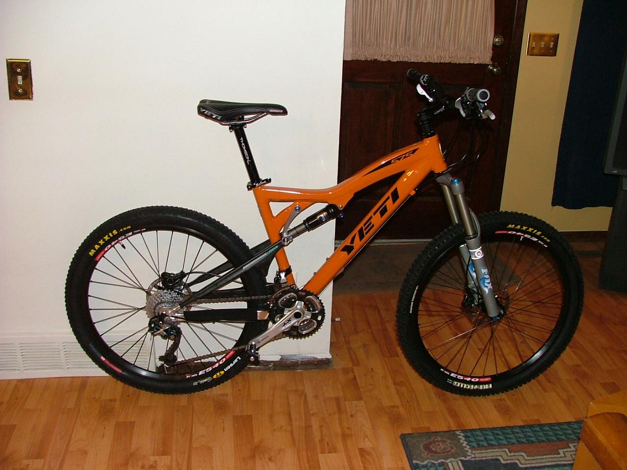 Yeti 575: A bright orange mountain bike leaning against a wall in an indoor setting. The bike features a sleek frame design, front suspension, and thick tires with visible tread patterns. The background includes a wooden door and a patterned rug on the floor.