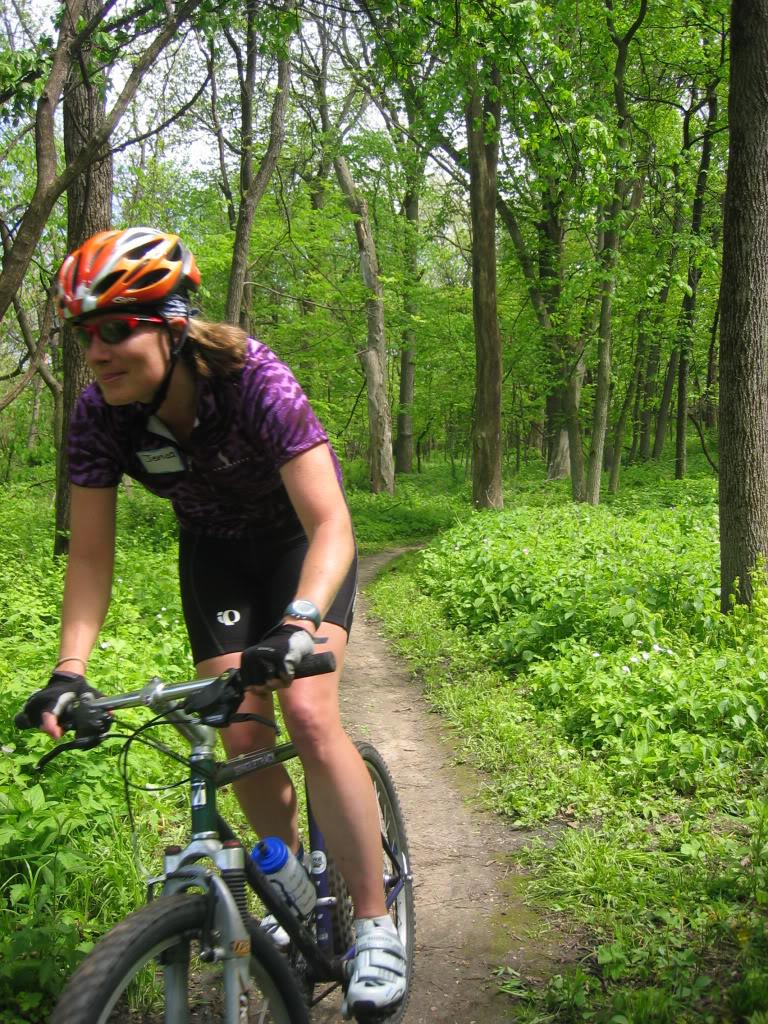 A cyclist wearing a helmet and sunglasses rides a mountain bike along a dirt trail in a lush green forest, surrounded by trees and vibrant foliage.