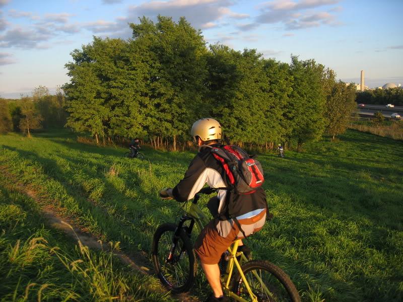 A cyclist wearing a helmet and a backpack is riding down a grassy hill, with trees in the background under a blue sky. Another cyclist can be seen further down the hill, while a road runs alongside the grassy area. The scene is illuminated by warm evening light, creating a vibrant outdoor atmosphere.