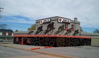 A building featuring a colorful mural depicting silhouettes of cyclists, with "Gung Ho" prominently displayed on the upper part. The design includes circular patterns reminiscent of bike wheels, set against a gray sky. The surrounding area appears suburban, with additional buildings in the background.