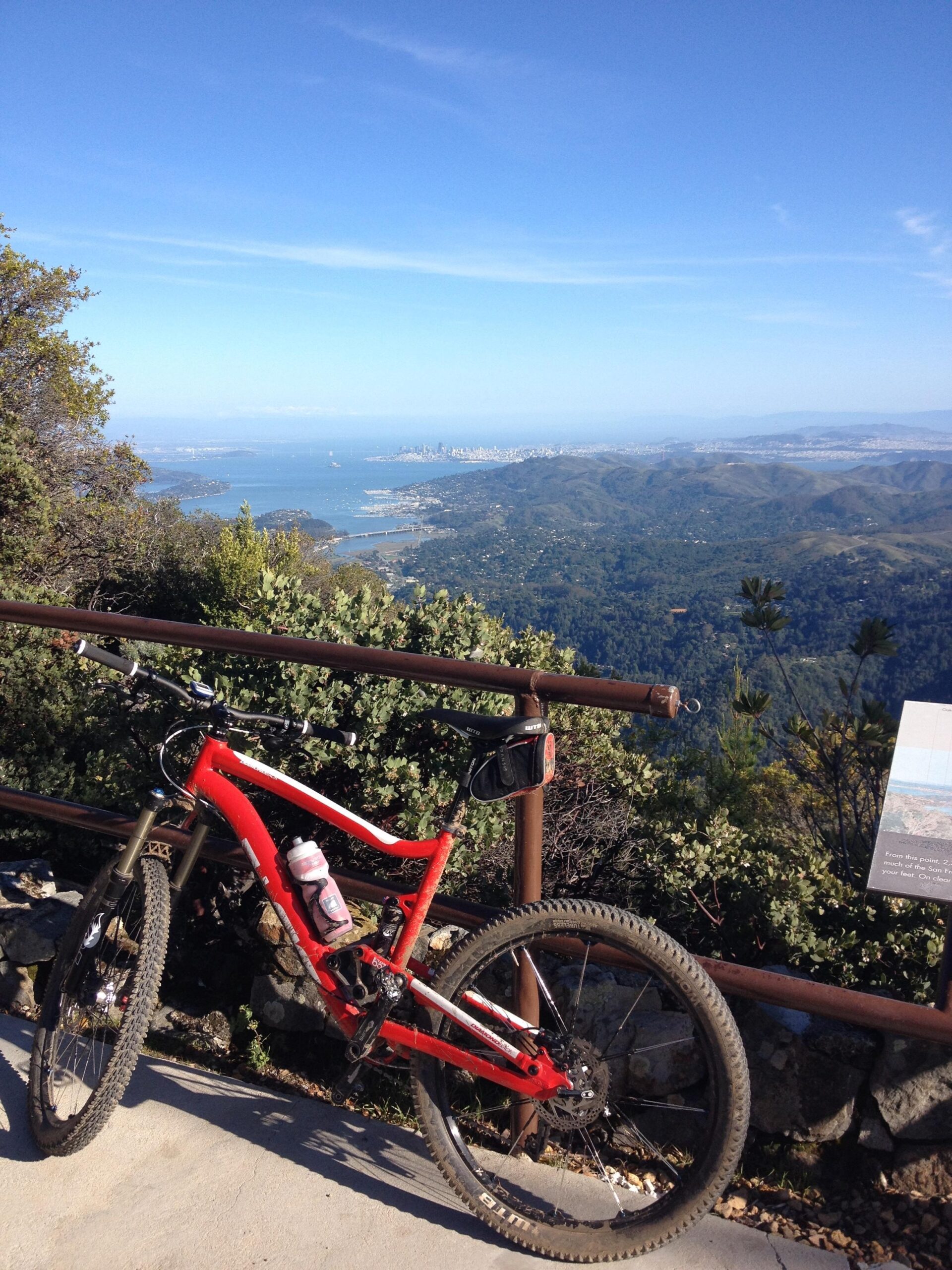 Diamondback Sortie Comp: A red mountain bike parked at a scenic overlook, with a panoramic view of a bay and city skyline in the background. Lush greenery surrounds the bike, and a metal railing borders the viewpoint. A water bottle is attached to the bike, and a informational sign is visible nearby. The sky is clear and blue, suggesting a sunny day.