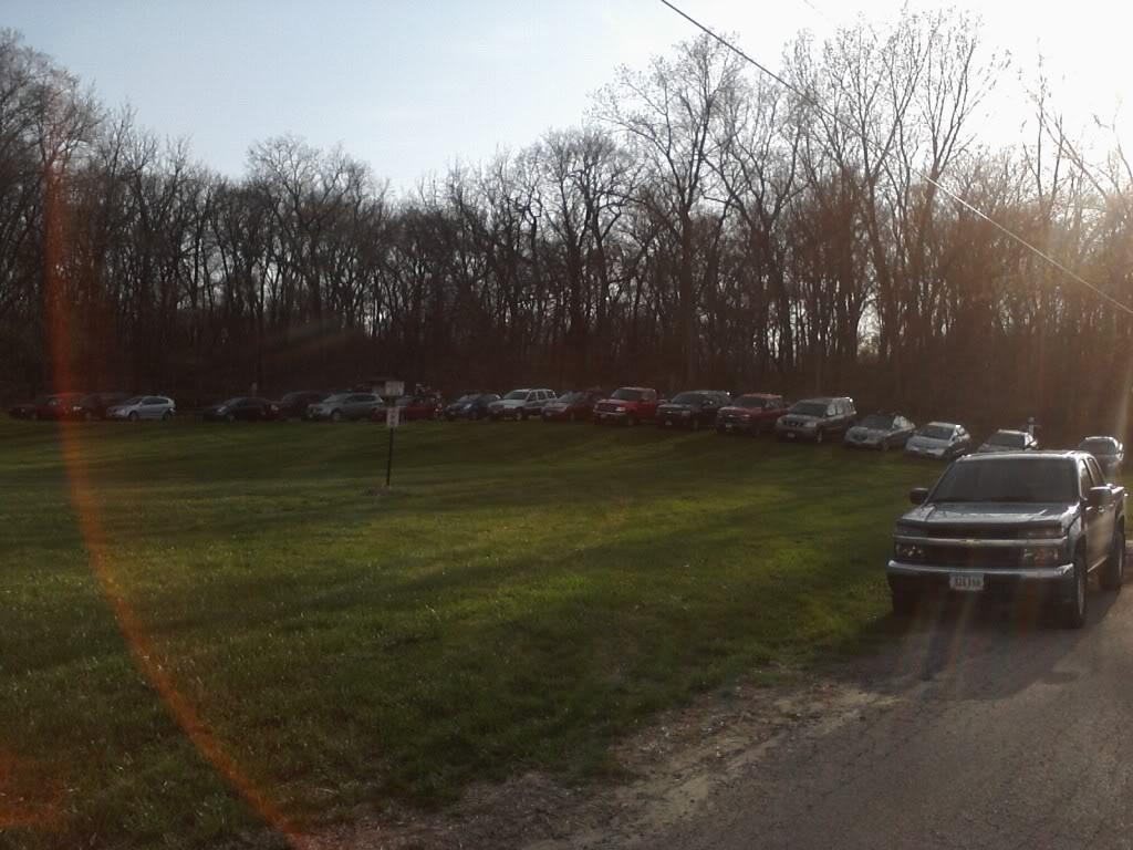 A row of parked cars along a grassy area, with trees in the background. The setting is likely an outdoor location, possibly a park or event venue. The foreground features a black pickup truck, and additional vehicles are arranged on the grass behind it. The time of day appears to be late afternoon or early evening, with soft sunlight illuminating the scene.