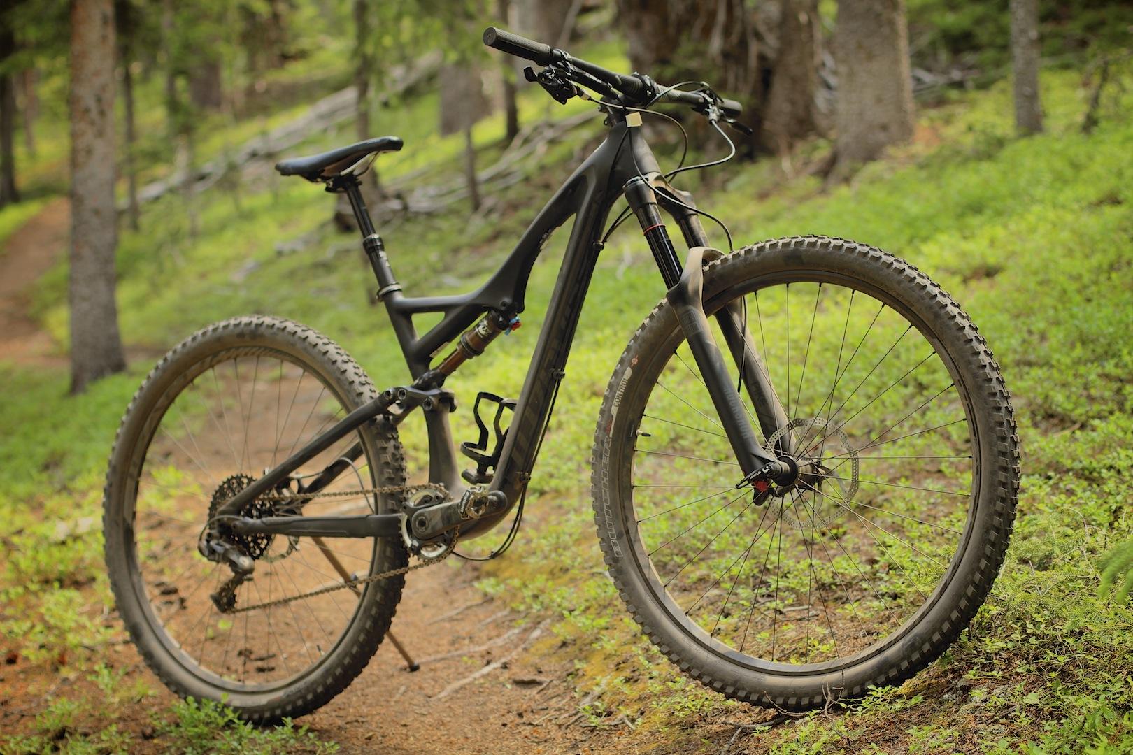 Specialized Stumpjumper FSR: A mountain bike is positioned on a dirt path surrounded by lush greenery and trees. The bike features a sleek black frame, thick tires with prominent treads, and visible suspension components. The background shows a serene forest setting with soft, diffused light filtering through the trees.