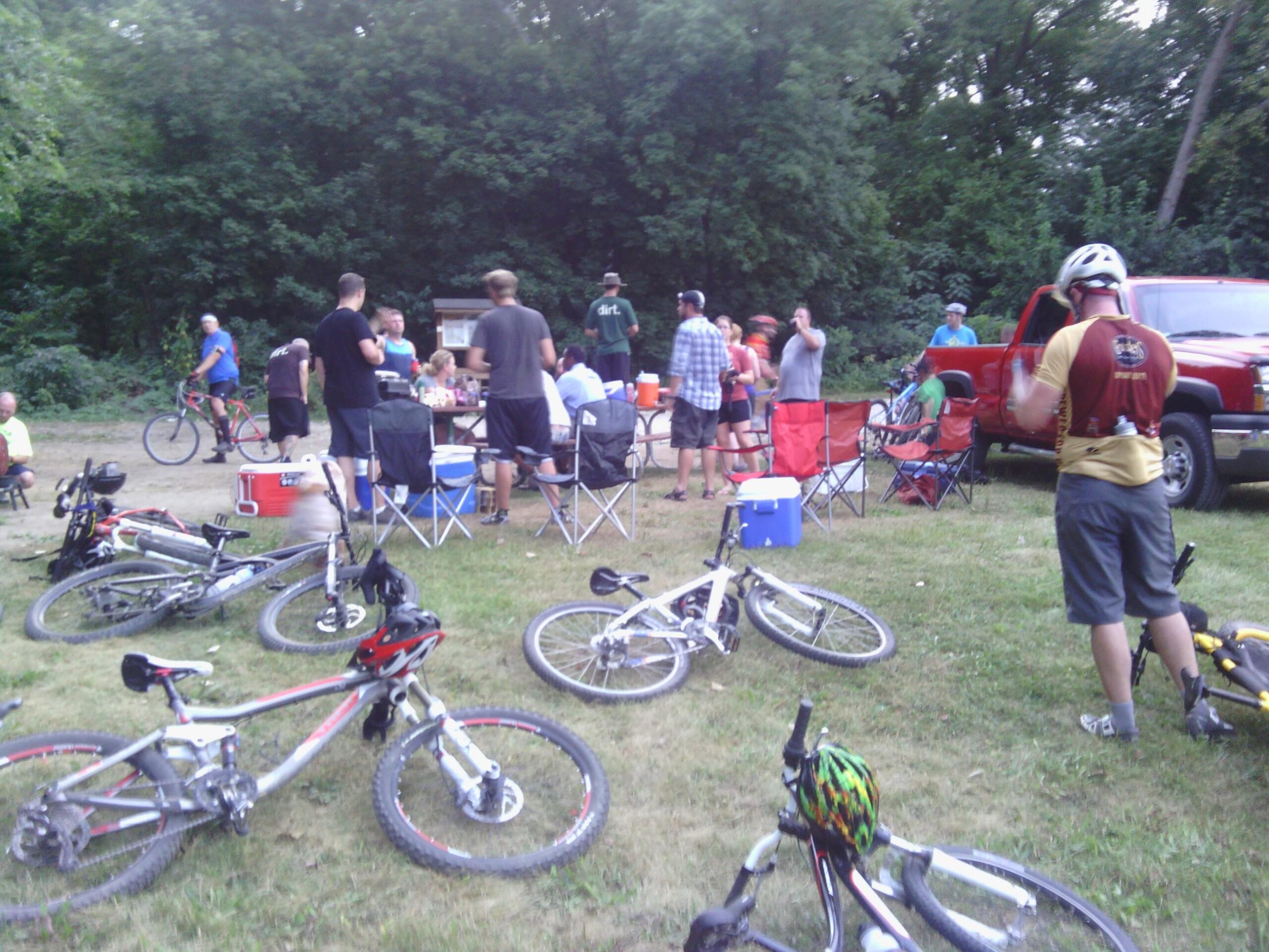 A group of people gathered outdoors near their bicycles, enjoying a social event. Some individuals are seated around folding chairs while others are mingling and chatting. There are coolers and a red pickup truck parked nearby, surrounded by trees in the background. The scene captures a lively atmosphere of camaraderie among cyclists.