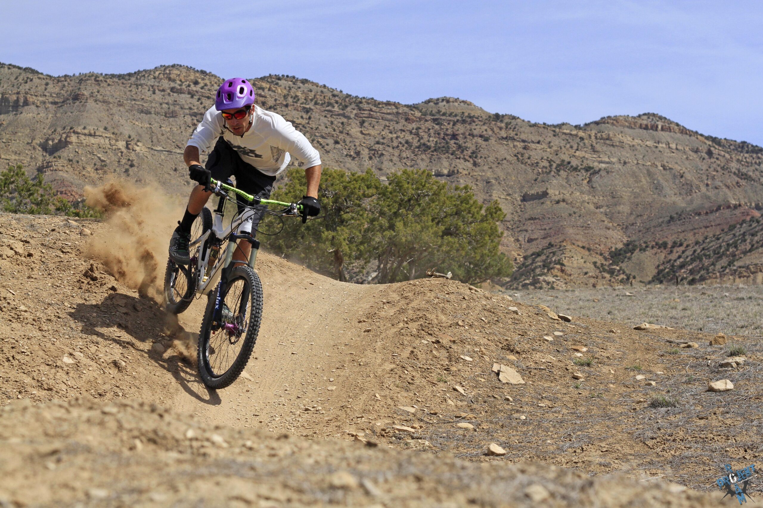 A mountain biker in a purple helmet and sunglasses rides a dirt track, kicking up dust as he navigates a turn. The background features rocky hills and sparse vegetation under a clear sky, showcasing an outdoor biking adventure. 18 Road Trails / North Fruita Desert mountain bike trail.