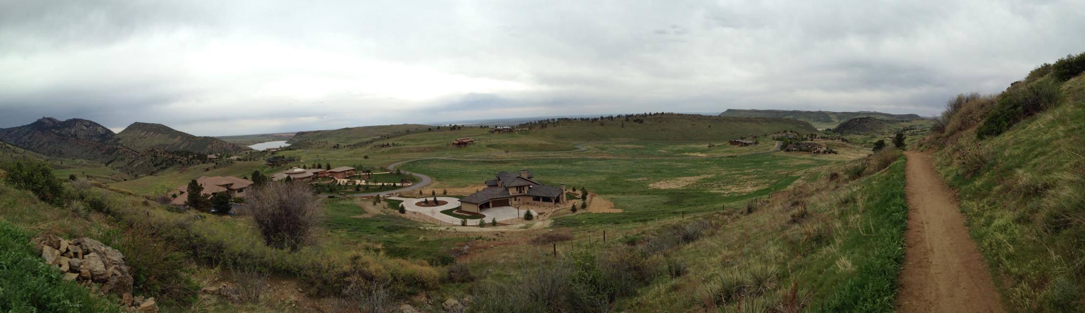 A panoramic view of a lush green landscape featuring rolling hills, low mountains, and a winding path leading into the distance. In the foreground, there are several modern homes nestled among the greenery, while the background showcases a lake and a cloudy sky. The scene captures the beauty of nature and serene suburban living. White Ranch mountain bike trail.