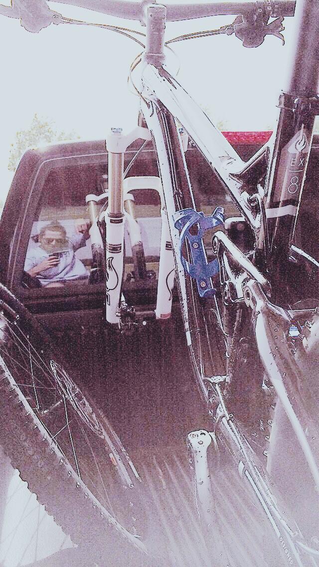 Trek Fuel EX 8: A bicycle with a sleek design is secured in the back of a pickup truck, with its front fork visible. In the reflection of the truck window, a person is seen taking a selfie, holding a phone. The background features greenery, suggesting an outdoor setting.