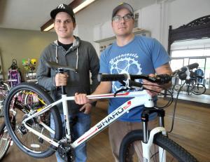 Two men stand in a bicycle shop, smiling as they pose with a mountain bike. One man is holding the bike by the handlebars, while the other stands beside him, wearing a hoodie and a baseball cap. The shop interior features various bicycles and biking gear in the background.