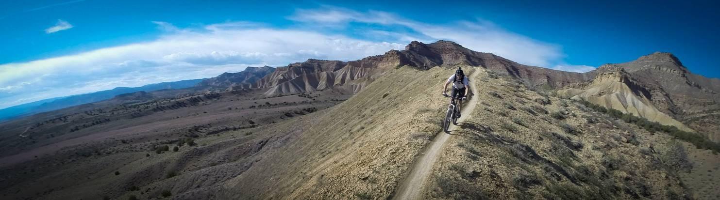 A mountain biker rides along a narrow trail on a hillside, surrounded by rugged terrain and distant mountains under a clear blue sky with wispy clouds. Zippety Do Dah mountain bike trail.