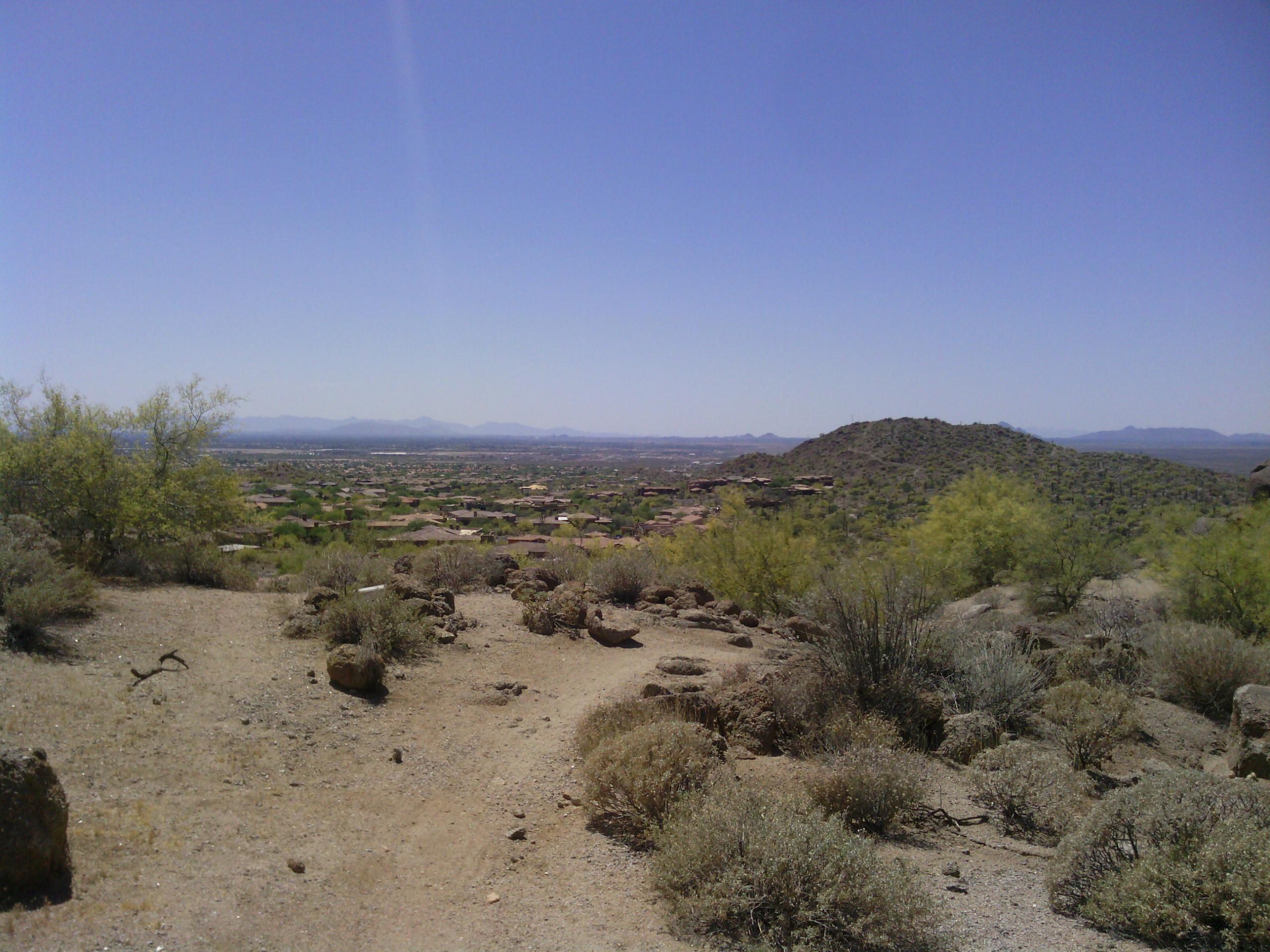 A panoramic view of a southwestern landscape featuring sparse vegetation, rocky terrain, and hills in the distance under a clear blue sky. The foreground includes a dirt path through desert shrubs, with scattered rocks, leading towards a valley filled with homes and further mountains in the background.