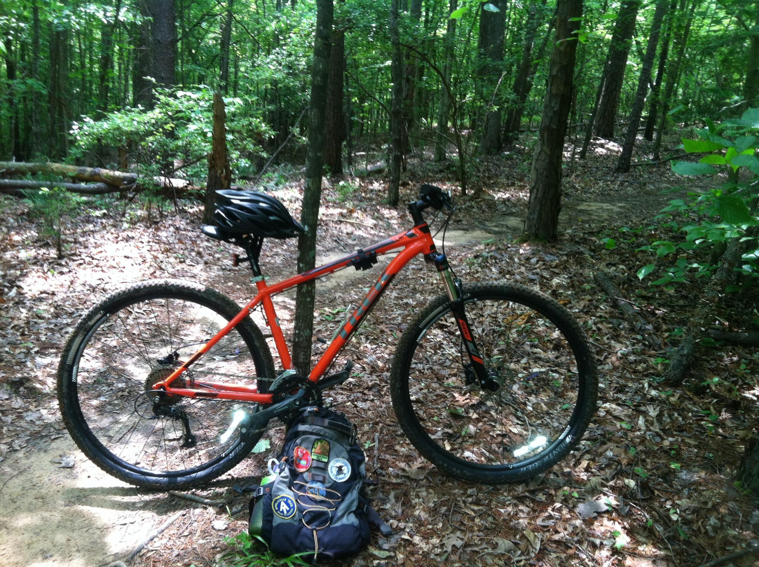 Trek X-Caliber 7: A bright orange mountain bike resting on a dirt path surrounded by green trees and foliage in a wooded area. A black helmet is secured on the bike's seat, and a backpack with various patches sits on the ground nearby, with fallen leaves scattered around.