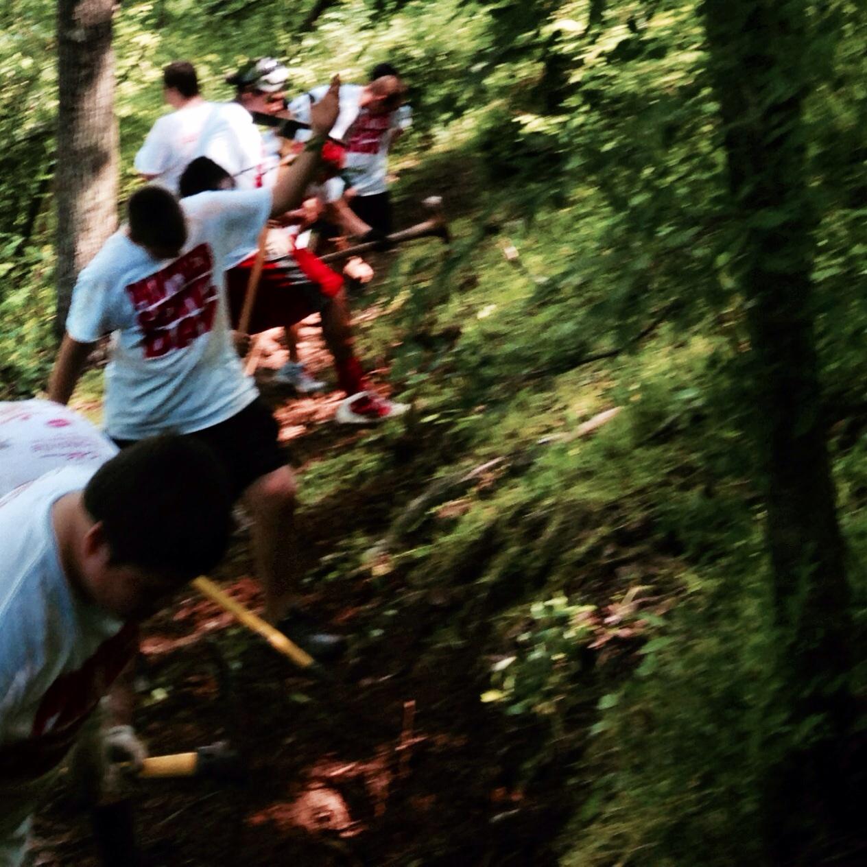 A group of volunteers wearing matching white t-shirts is working together on a trail, using shovels and tools to clear the area. The scene is set in a lush, green forest with trees and underbrush surrounding them, showcasing an outdoor service or community project. Spadra Creek Nature Trail mountain bike trail.