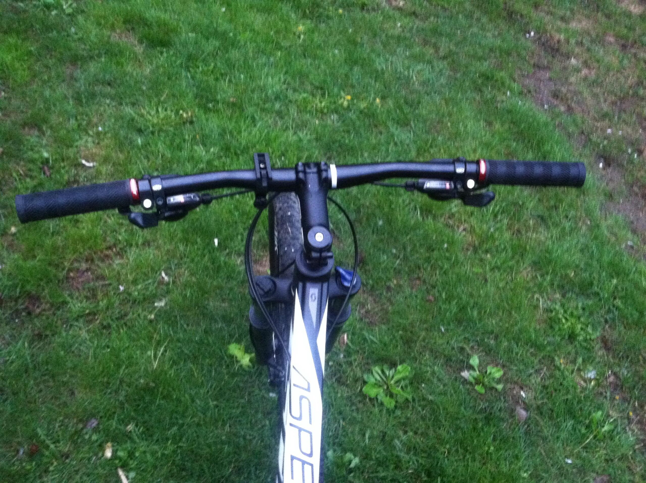 Scott aspect 940: A top view of a mountain bike's handlebars, featuring black grips and control levers, set against a green grassy background. The bike frame is partially visible, displaying the word "ASPEN" in a distinctive font.