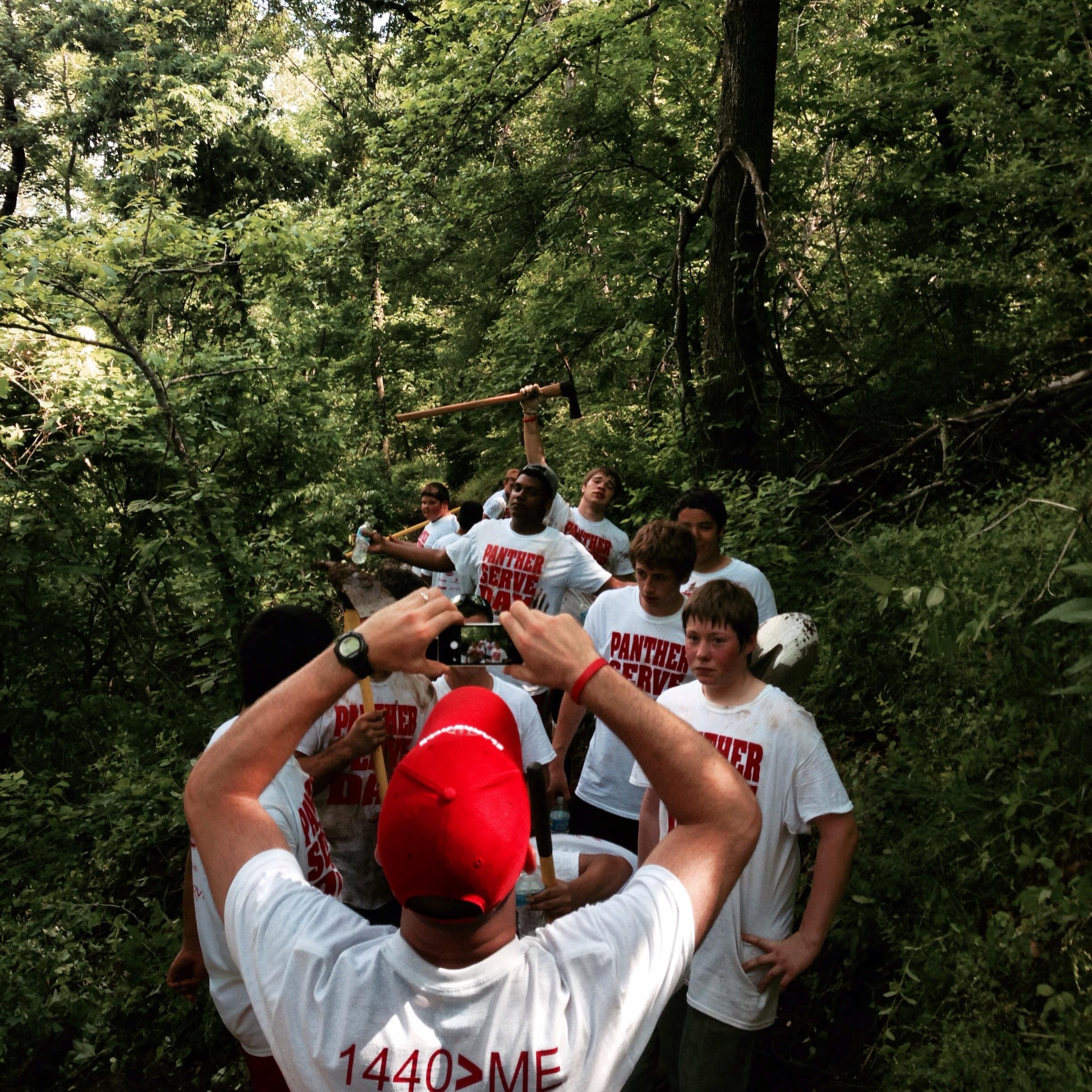 A group of young volunteers in matching white T-shirts with "PANTHER SERVE" printed on them pose for a photo in a wooded area. One person is holding a handheld tool, and others are smiling and holding water bottles. The scene is lively, showcasing teamwork and outdoor community service. Spadra Creek Nature Trail mountain bike trail.