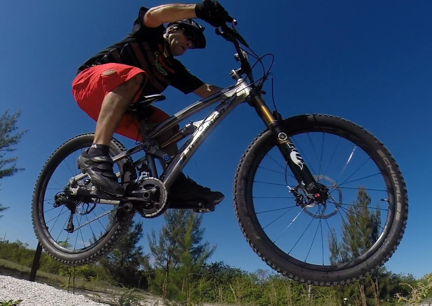 Transition Covert: A cyclist performing a jump on a mountain bike against a clear blue sky. The rider is wearing a black helmet and gloves, with a black and red outfit. The bike features large tires with visible tread, and the background includes green trees and shrubs.
