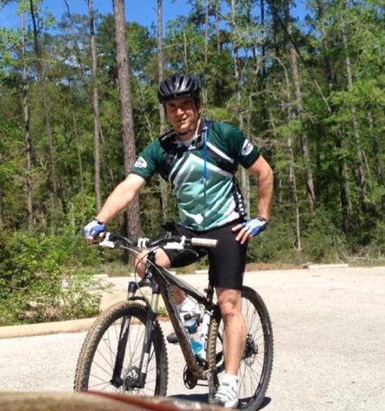 Specialized Rockhopper Comp 29: A cyclist in a green and black jersey poses on a mountain bike in a wooded area, surrounded by trees under a clear blue sky. The cyclist is wearing a helmet and gloves, with one hand on the handlebars and a confident smile.