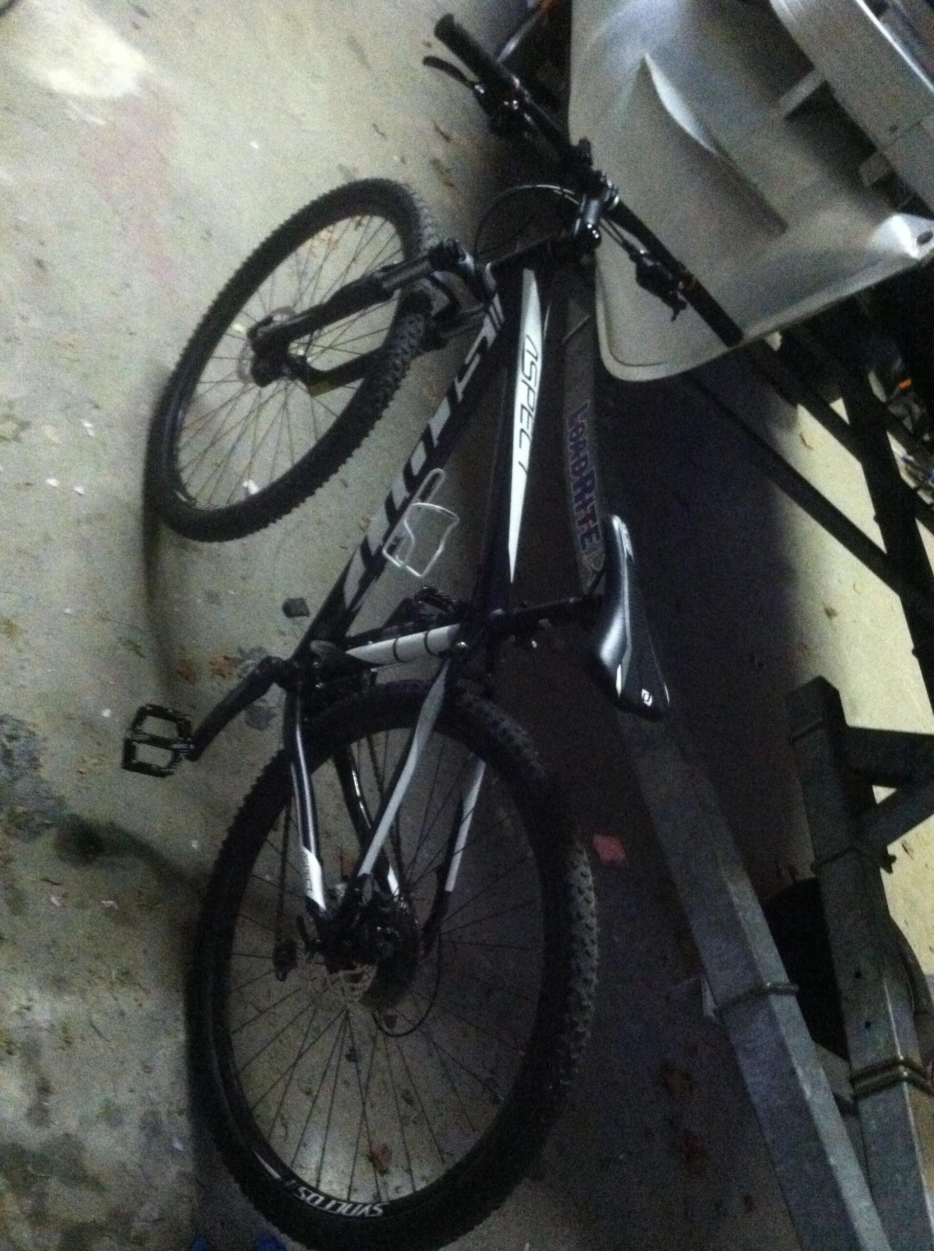 Scott aspect 940: Mountain bike resting on a concrete surface, angled to show its frame and wheels. The bike features a black and white color scheme, with visible components like the handlebars, seat, and pedals. Background elements include a partially visible trailer and debris on the ground, indicating an indoor or garage setting.