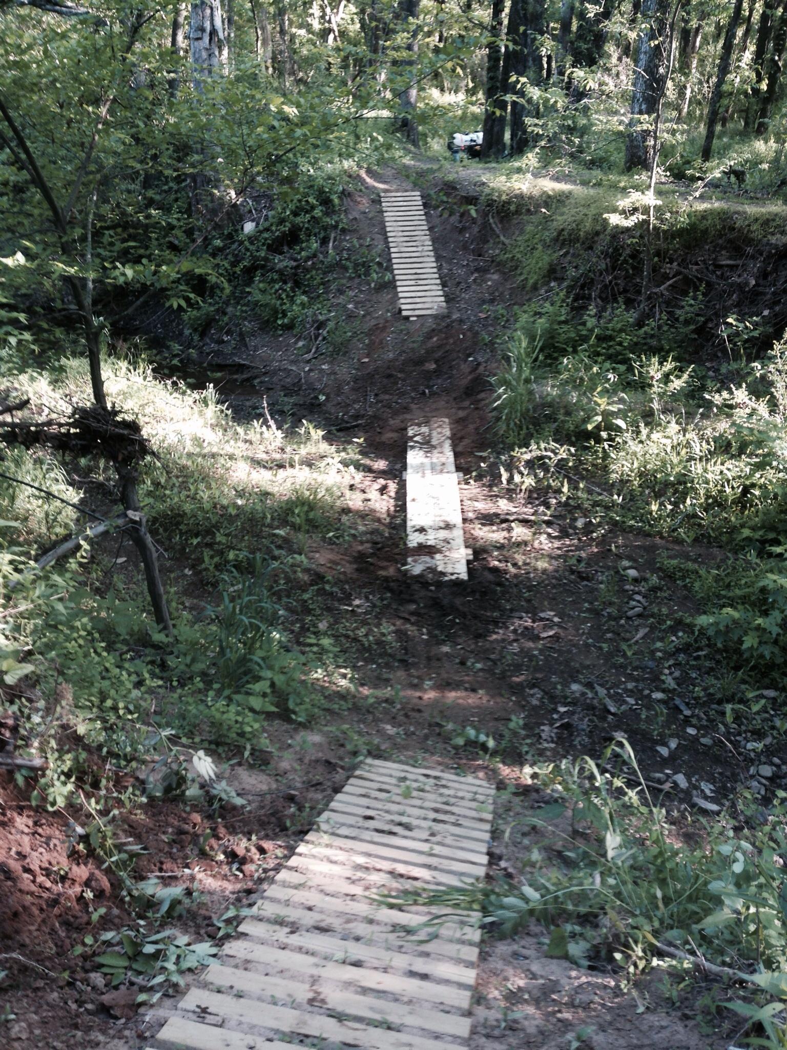 A narrow wooden path crossing a small ravine in a forested area, surrounded by greenery and trees. The path appears rustic, leading from one side of the ravine to the other, with natural foliage on both sides. Spadra Creek Nature Trail mountain bike trail.