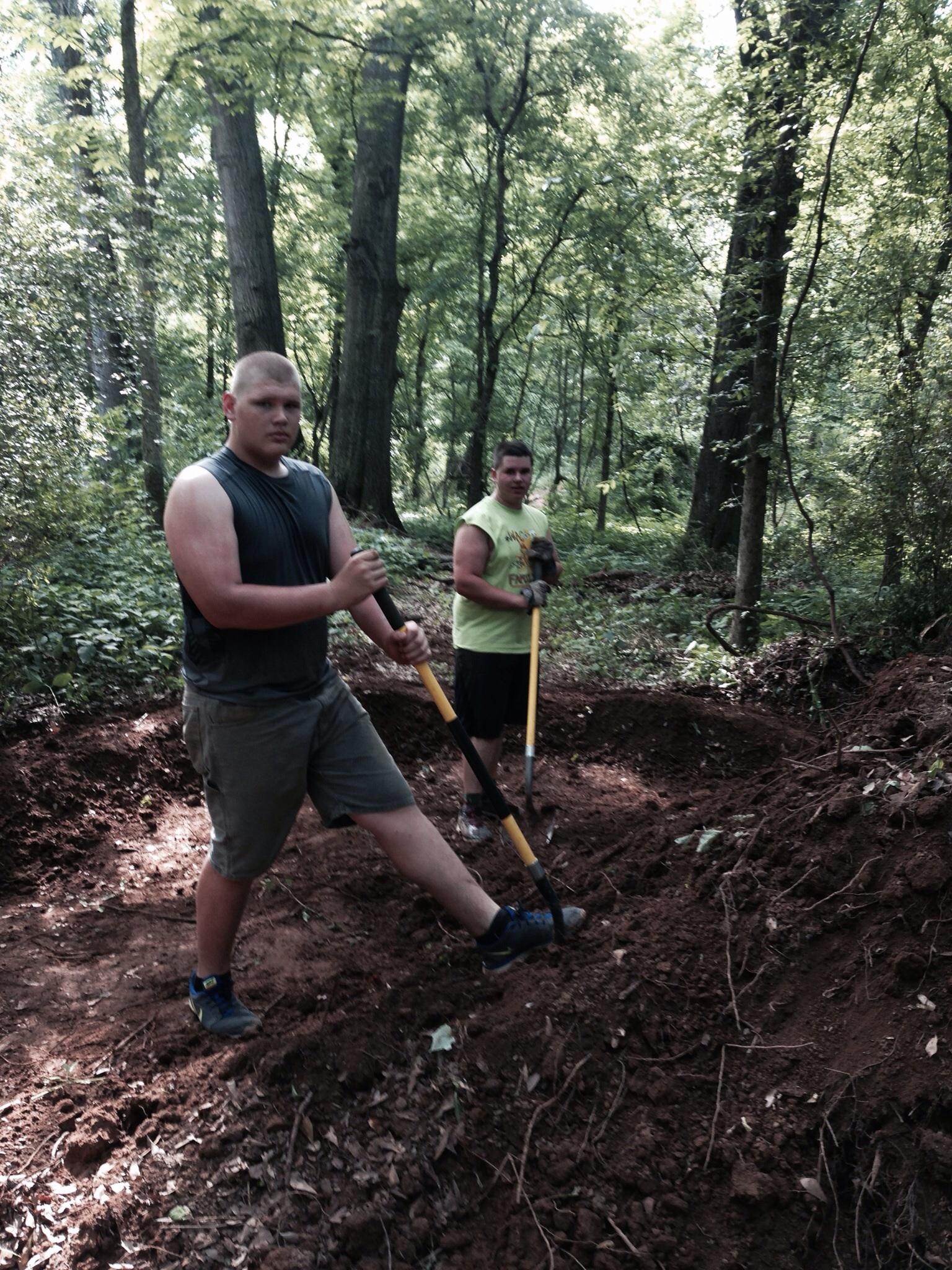Two young men working in a wooded area, using shovels to dig in the dirt. One is wearing a sleeveless shirt and has a serious expression, while the other, in a bright yellow shirt, stands nearby holding a shovel. The scene is surrounded by trees and greenery, indicating an outdoor project. Spadra Creek Nature Trail mountain bike trail.