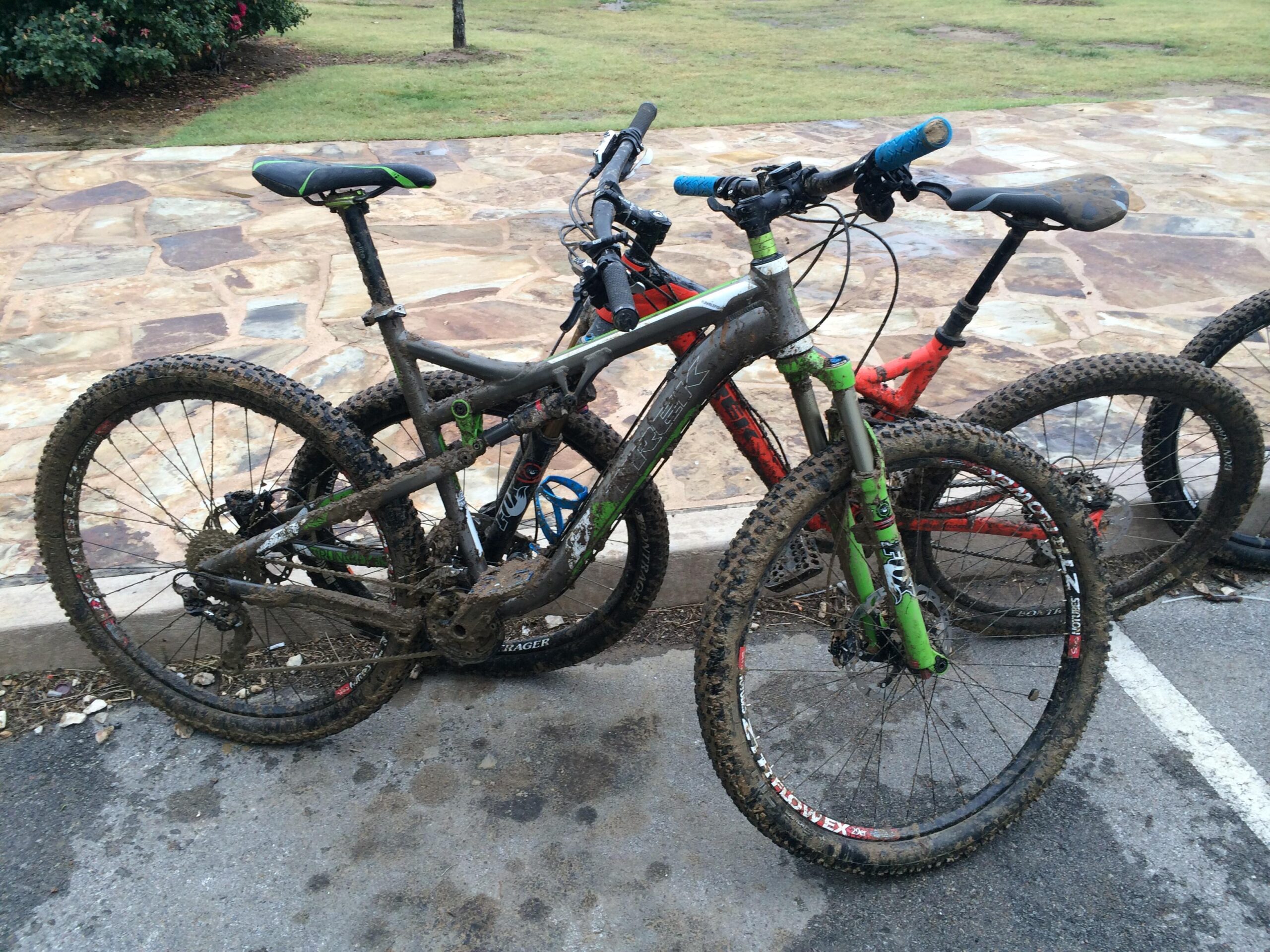 Trek Rumblefish: Two mountain bikes are leaned against a stone pathway, both covered in mud after a ride. One bike is primarily gray with green accents, while the other is red. The background features grassy areas and trees.