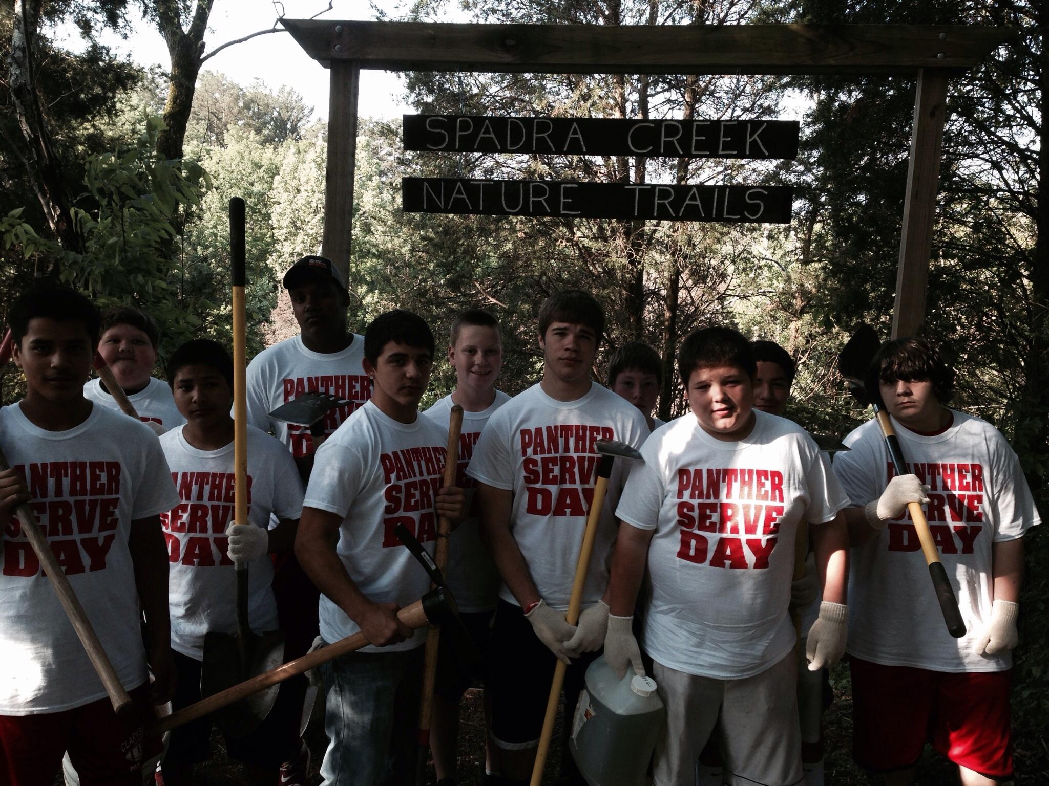 A group of young volunteers stands in front of a wooden sign that reads "Spadra Creek Nature Trails." They are wearing matching white T-shirts with "PANTHER SERVE DAY" printed in red. Each person holds a tool, such as a shovel or hoe, as they prepare to engage in community service. Lush greenery surrounds them, indicating a natural outdoor setting. Spadra Creek Nature Trail mountain bike trail.