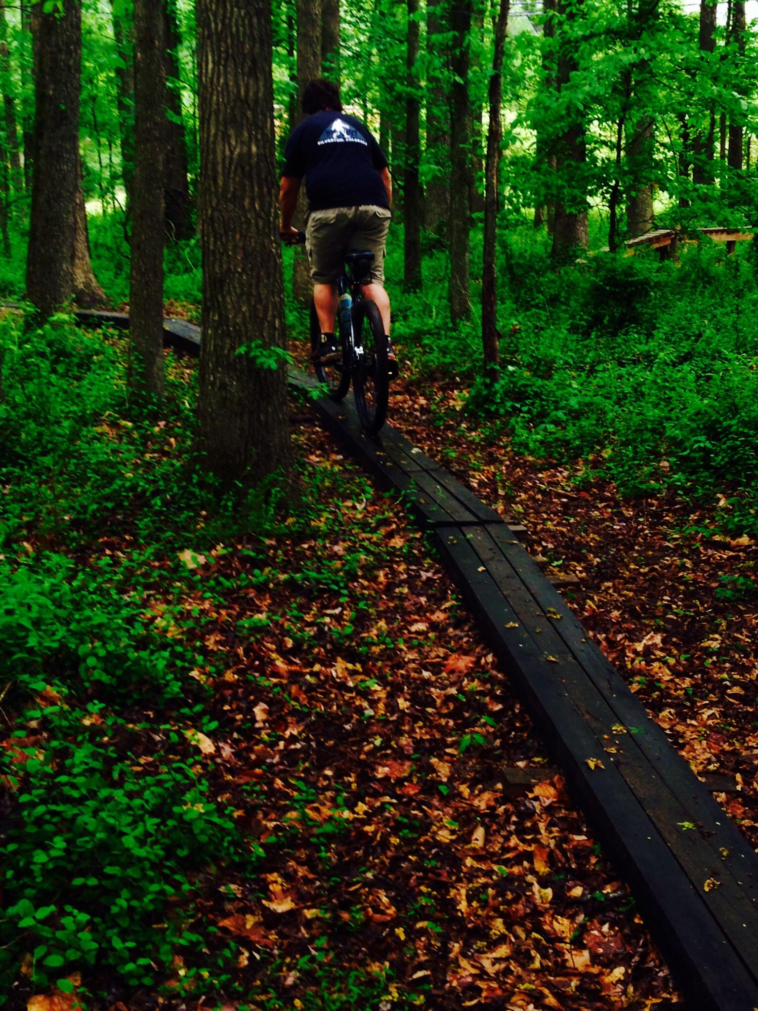 A person riding a bicycle on a narrow wooden path through a dense forest, surrounded by lush green trees and scattered autumn leaves on the ground. Spadra Creek Nature Trail mountain bike trail.