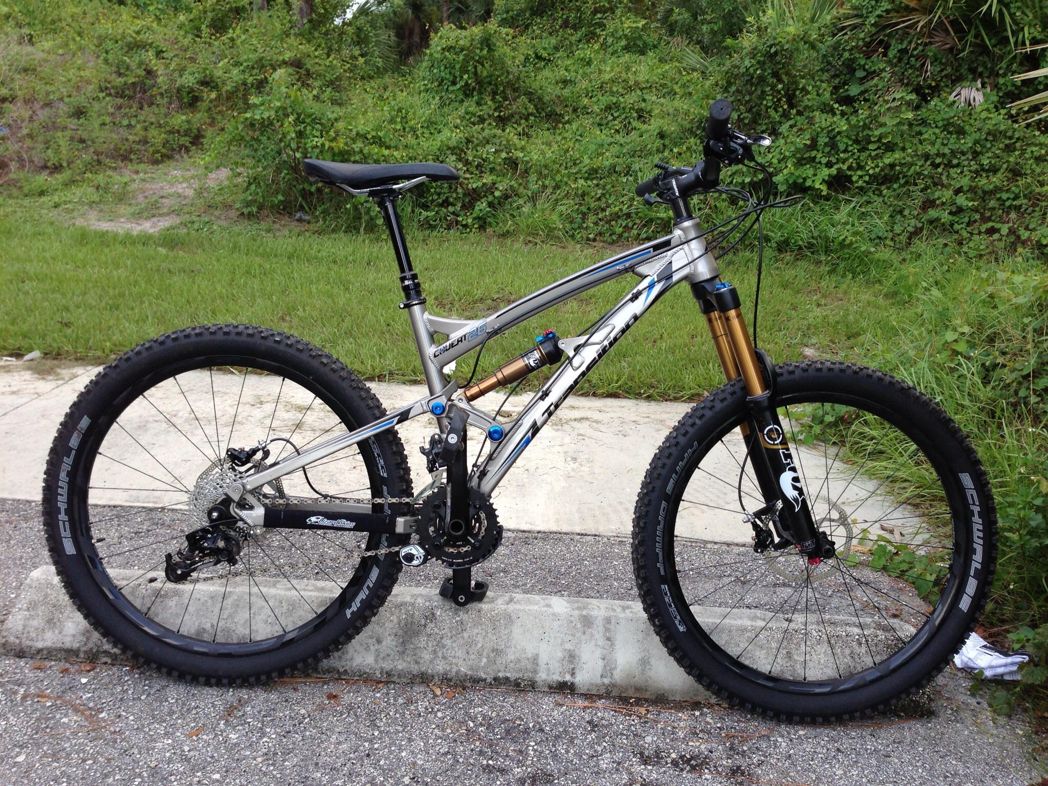Transition Covert: Mountain bike parked on a concrete curb, featuring a silver frame with blue and gold accents. The bike has thick, knobby tires designed for off-road terrain, a prominent front suspension fork, and a visible drivetrain with gears and a rear derailleur. Lush green grass and foliage are in the background.
