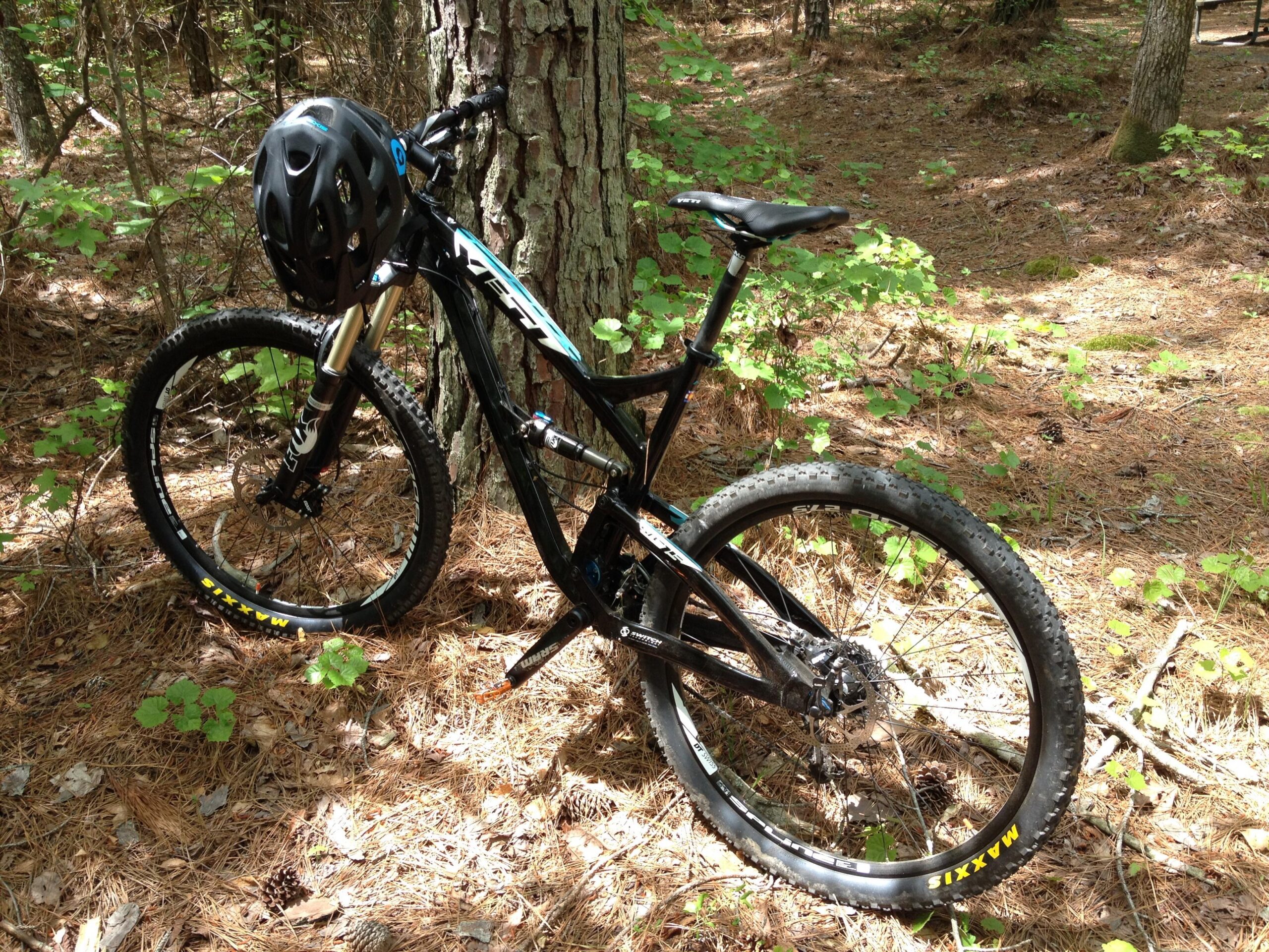 Yeti SB-75: A mountain bike with a black frame and yellow Maxxis tires resting against a tree in a forested area, surrounded by pine needles and greenery. A black helmet is placed on the handlebars, indicating a setup for biking in nature.
