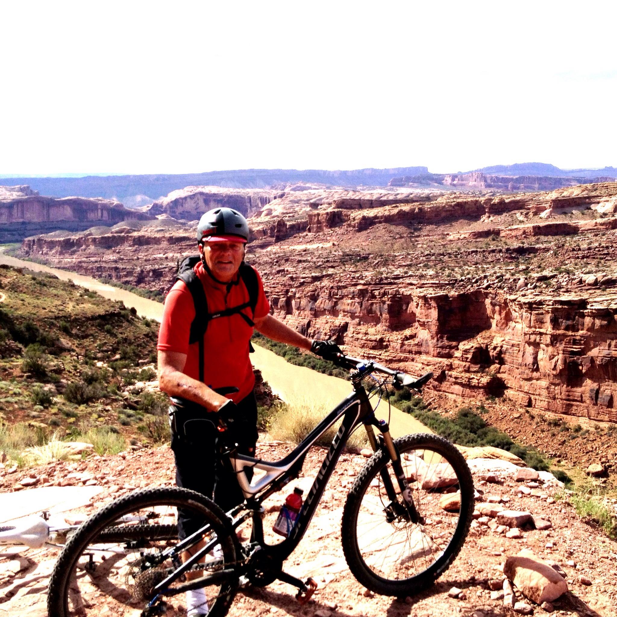 Specialized Stumpjumper FSR Comp 29: An older man in a red shirt and helmet stands next to a mountain bike on a rocky ledge, overlooking a deep canyon. The landscape features steep cliffs and a winding river below, with a bright sky in the background.