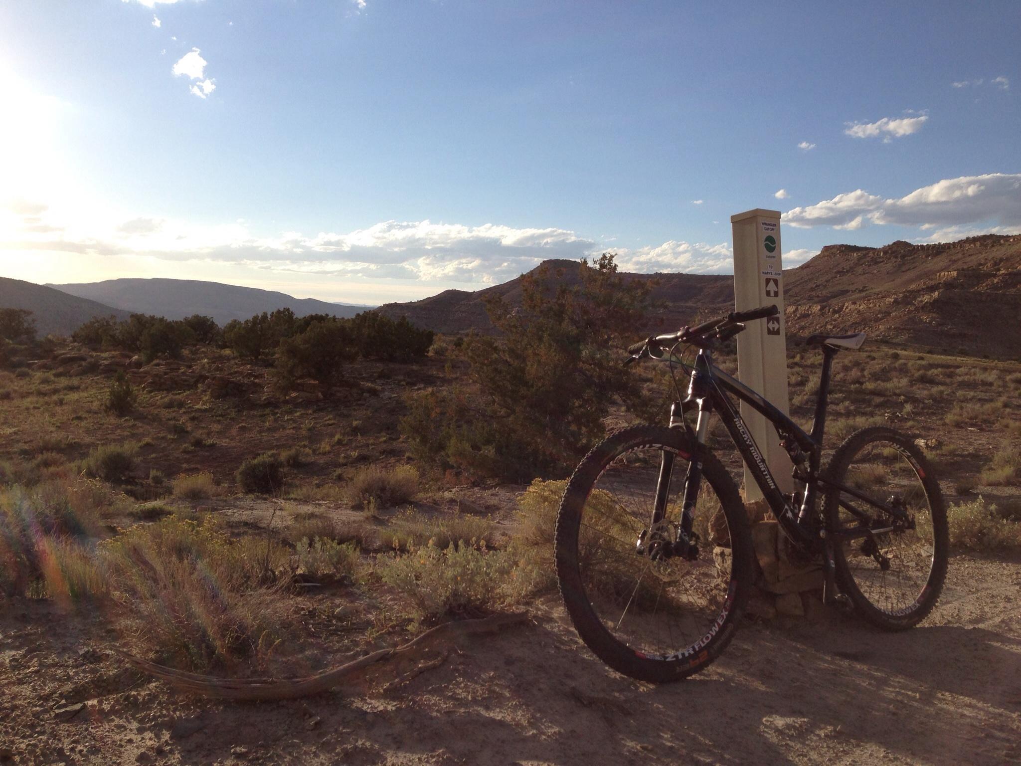 A mountain bike leaning against a trail marker in a desert landscape, with rolling hills and shrubs in the background under a clear sky. The sun is setting, casting warm light across the scene. Mary's Loop / Horsethief Bench mountain bike trail.